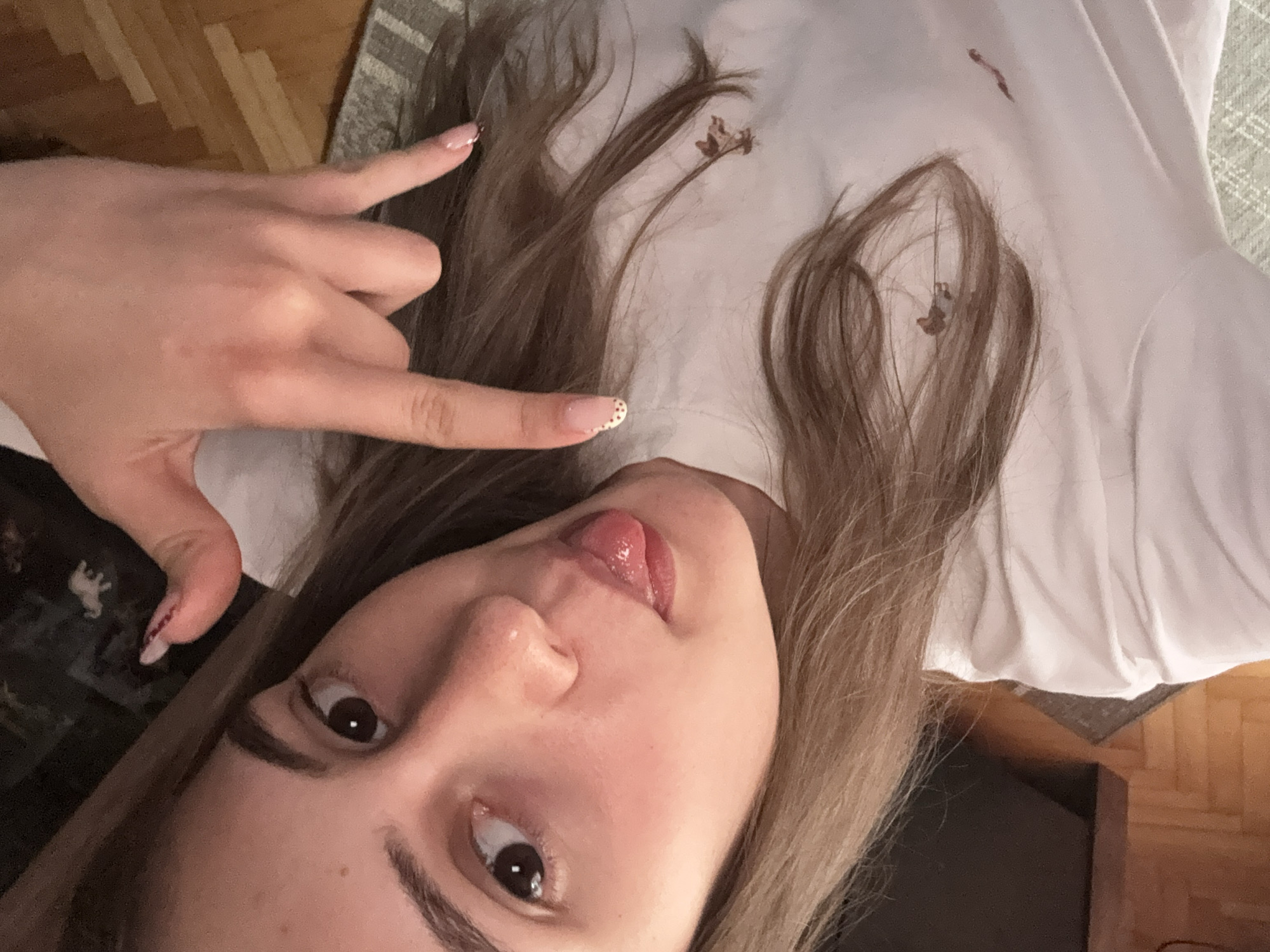 This rotated close-up image depicts a young female indoors, possibly in Panagyurishte, Bulgaria. The subject is seen from a low, oblique angle, suggesting she might be lying down or holding the camera at an unusual orientation. She has long, light brown hair, which is spread out and partially covers a white top she is wearing. Small pieces of dried plant matter or debris are visible on her hair and the white fabric.
Her face is prominent, featuring dark brown eyes and well-defined, dark eyebrows. She displays a playful expression, sticking her tongue out slightly. Her left hand is raised, making a gesture commonly known as the "rock on" or "devil horns" sign, with the index and pinky fingers extended. Her nails are manicured in a French tip style, with additional small red dots or patterns visible on the tips of some nails, particularly her index finger.
The background indicates an indoor setting, with a light wooden floor visible in parts of the frame, alongside a partially seen light-colored, textured rug or carpet. The image conveys a casual and direct engagement with the camera, characteristic of a selfie. There is no visible text in the image.