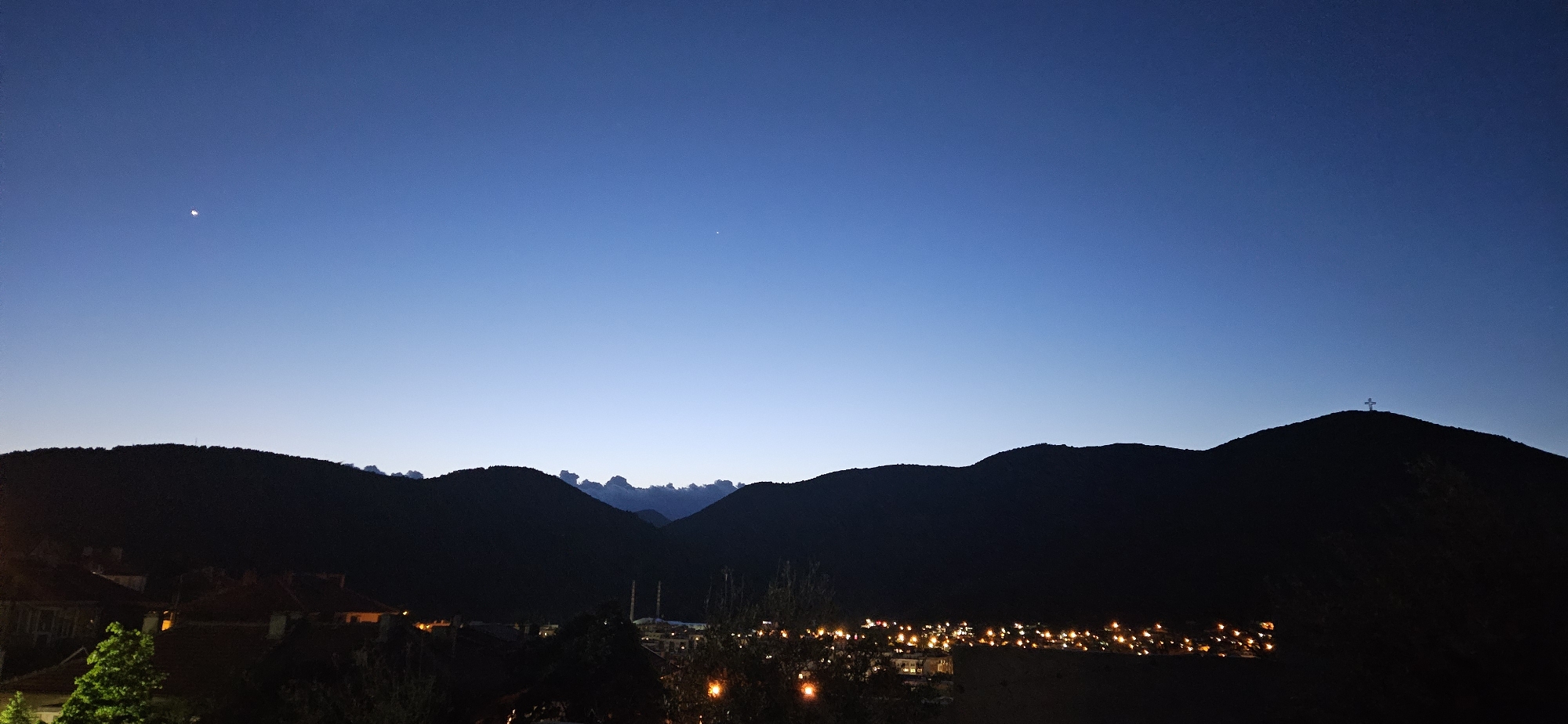 The image depicts a twilight scene in an urban area within a mountainous region. The sky transitions from a dark blue at the top to a lighter blue near the horizon. A bright celestial object is visible in the upper-left quadrant of the sky. Silhouetted mountain ranges extend across the midground. Below the mountains, the lights of a town or city are visible, with clusters of yellow lights indicating buildings and streets. In the foreground, there are indistinct shapes that appear to be trees and rooftops of buildings. A cross is visible atop a mountain peak on the right. The location is in the city of Unknown, Unknown.
