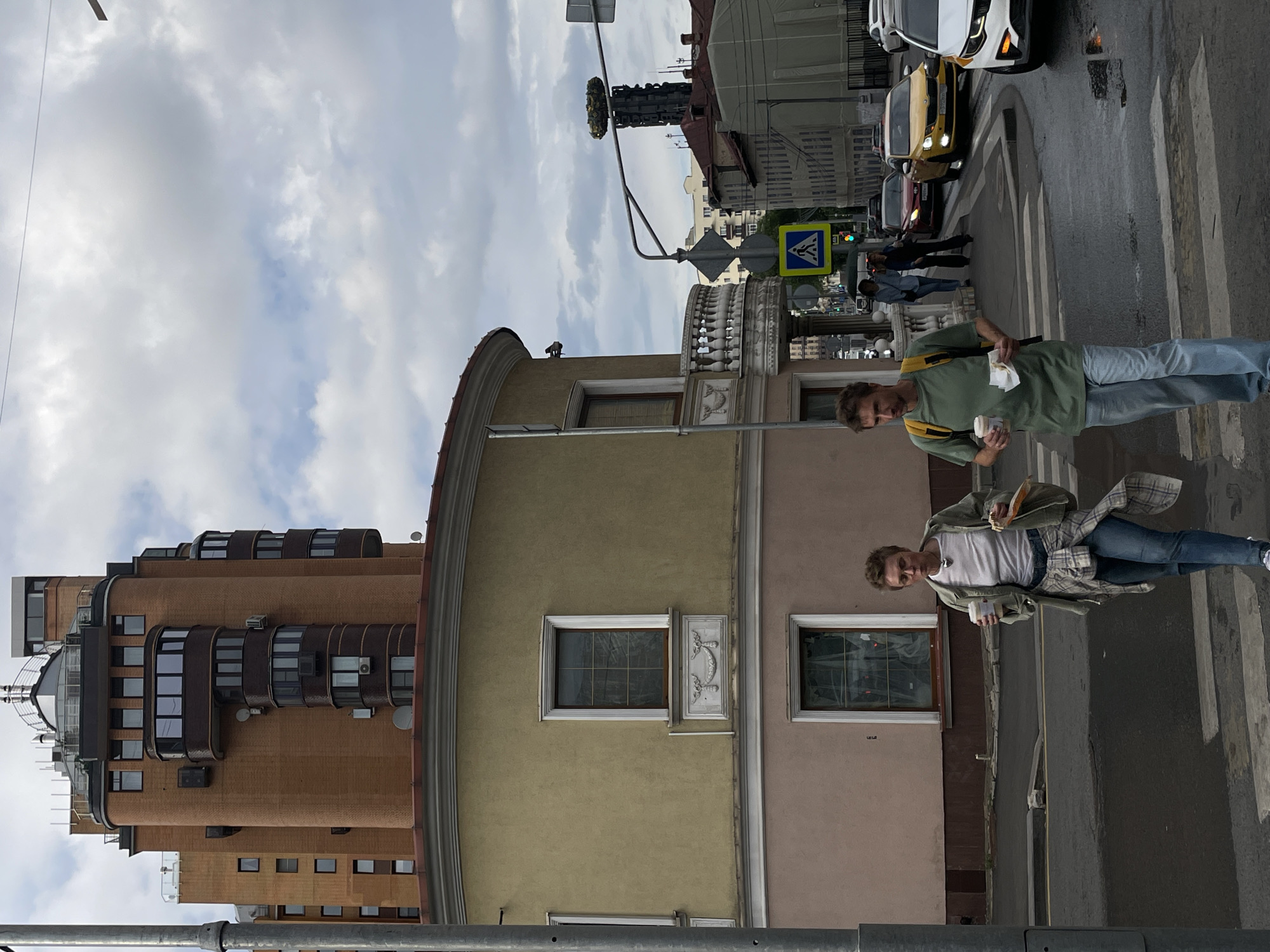 Two individuals are walking across a pedestrian crossing on a cloudy day in Moscow, Russia. The woman on the left, with short, cropped hair and glasses, wears a light olive-green jacket over a white t-shirt, blue jeans, and a plaid shirt tied around her waist. She holds a disposable coffee cup in her right hand and a small wrapped item in her left. The man on the right, wearing a green t-shirt, light blue jeans, and a yellow backpack, also carries a disposable coffee cup and a wrapped food item. They are walking towards the viewer.
Behind them, the cityscape features a distinctive curved building with a pale yellow facade on the lower floors and a tall, multi-tiered brick structure above. The brick building has several rounded balconies and numerous windows. Further in the background, other buildings, traffic lights, street signs, and vehicles, including a yellow taxi, are visible. The street appears wet, suggesting recent rain. A lamppost stands to the left of the frame, and overhead wires are present.