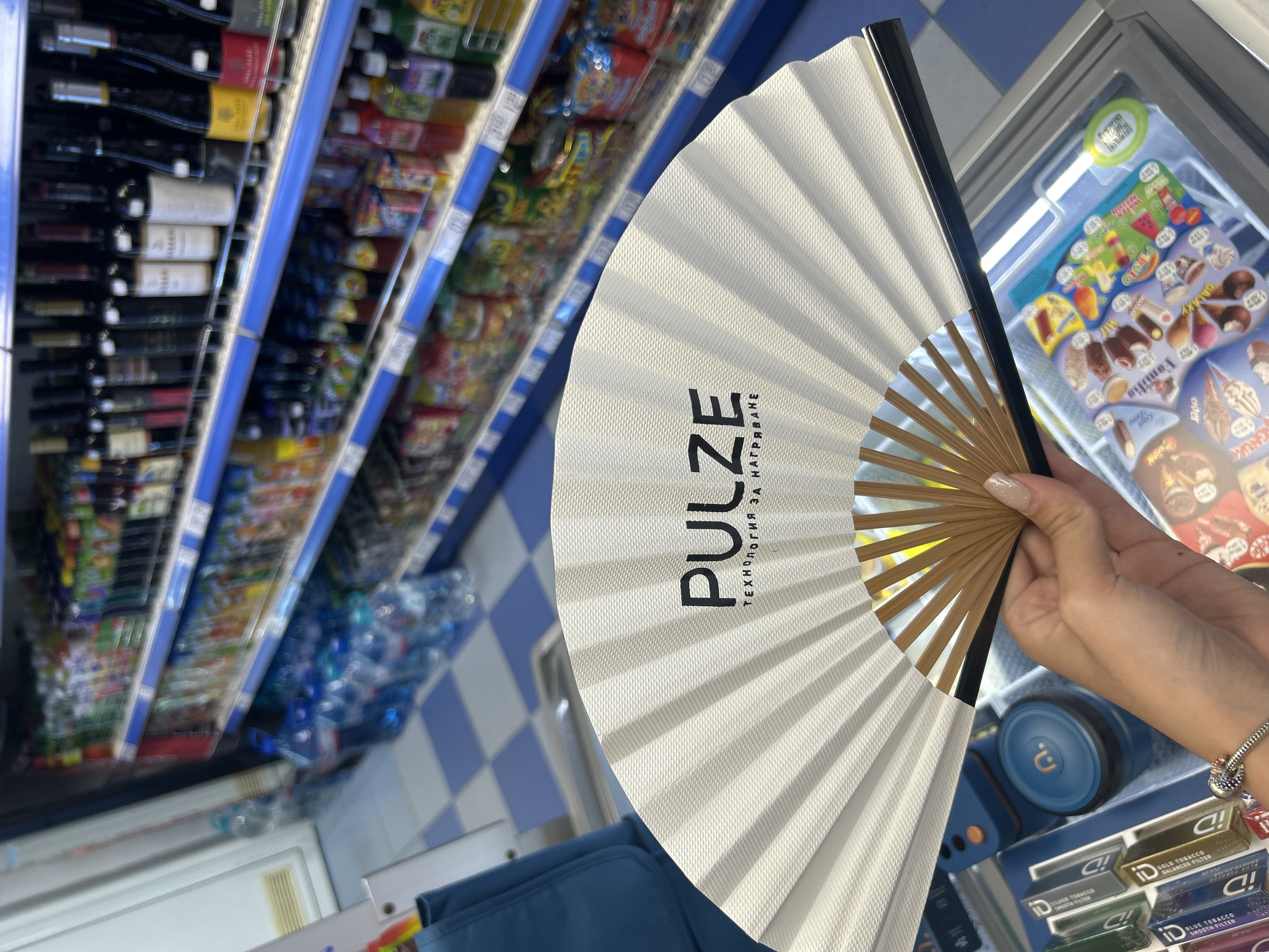 A person holds a white fan with a black border in a retail store. The fan is open, displaying the brand "PULZE" and Cyrillic text. In the background, shelves are stocked with various products, including bottles of water, wine, juices, and other items. The flooring is a checkered pattern of blue and white tiles. In the foreground, the edge of an open freezer containing ice cream is visible.