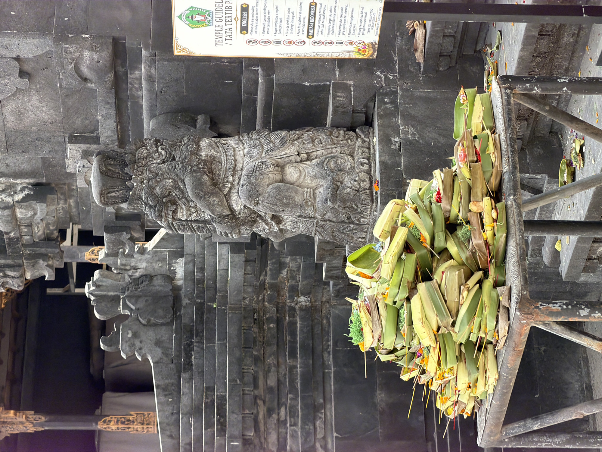 An outdoor stone structure with elaborate carvings features a stack of offerings in woven baskets made of leaves, placed on a metal table in front of a monumental stone statue. The offerings are adorned with yellow sticks and some green and red elements. To the right, a sign displays rules and information in English and Bahasa Indonesia for visitors to a temple. The background consists of grey stone stairs and walls, indicating a religious or historical site. The scene is likely located in Nusa Penida, Indonesia, a region known for its Hindu temples and cultural heritage.