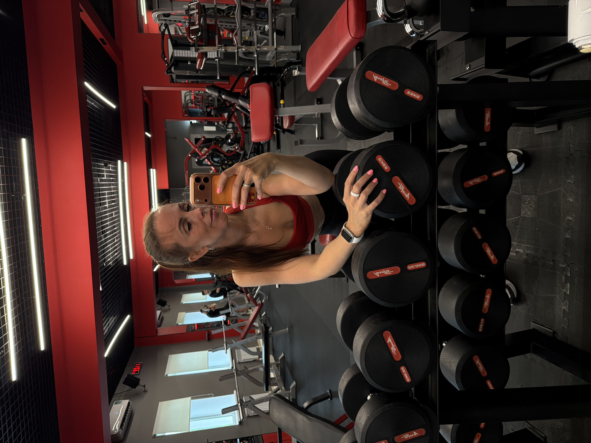 A woman is taking a selfie in a gym. She is wearing a red sports bra and black leggings, with her hair pulled back in a ponytail. She is holding a smartphone in her right hand, and her left arm is resting on a rack of dumbbells. The gym appears to be well-equipped, with various weight machines and other fitness equipment visible in the background. The lighting is bright, with fluorescent lights on the ceiling. The overall impression is of a modern and clean fitness facility. The location is a gym in Birobidzhan, Russia.