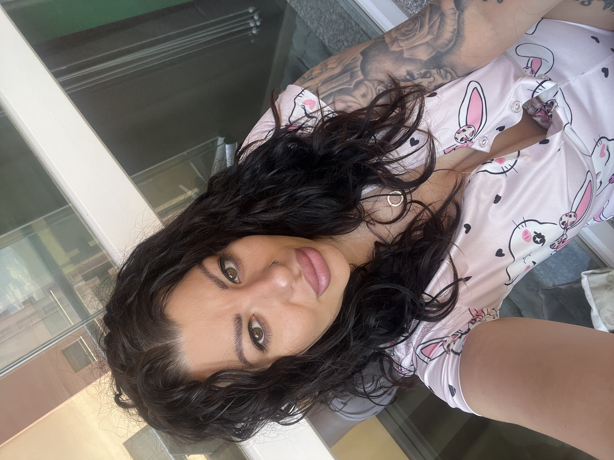A woman with dark curly hair and fair skin is taking a selfie indoors, presumably in a building in Plovdiv, Bulgaria, judging by the building visible through the window. She is wearing a pink pajama top with a rabbit pattern. She has a nose ring and a tattoo on her left arm. The background shows an exterior with windows and a building.