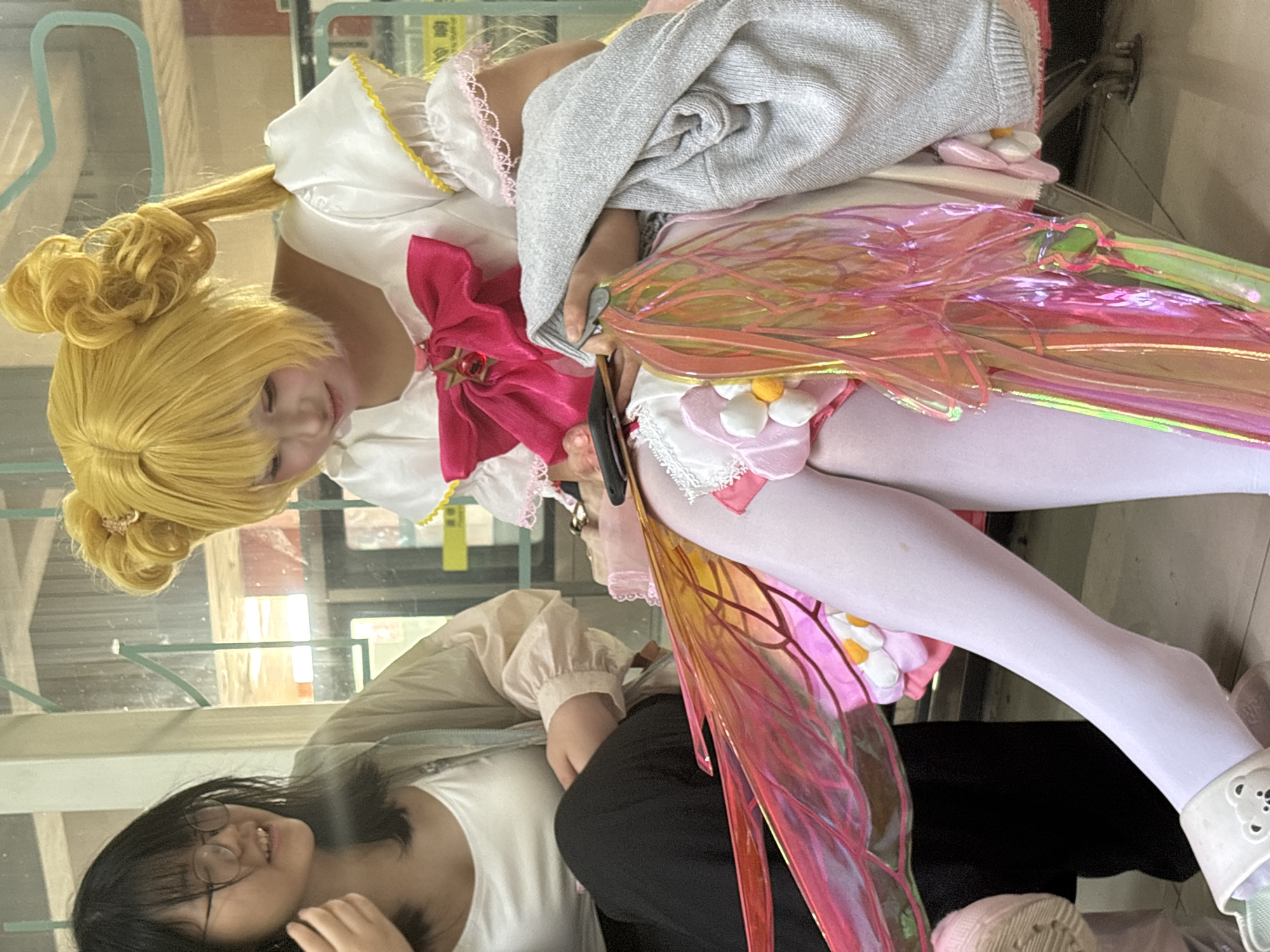 A woman dressed in a yellow wig and a pink and white costume with wings sits reflected in a glass surface across from another woman in glasses and a white shirt. The location is Yuzhong District, China.
