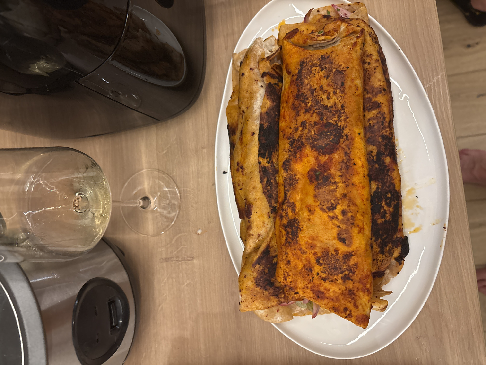 A close-up, overhead shot shows two golden-brown, rolled enchiladas on a white oval plate. The enchiladas appear to be filled with shredded meat and are topped with a vibrant orange sauce, some parts slightly charred from cooking. The plate rests on a light brown, wooden surface. To the upper left of the plate is a partial view of a metallic slow cooker with a black lid, and to the upper right, a black appliance, possibly an air fryer. Between these appliances and slightly behind the enchiladas is a wine glass filled with a pale yellow liquid, presumably white wine. The scene is lit by artificial light, casting subtle shadows. The background reveals a hint of a kitchen or dining area. The image was taken in Viernheim, Germany.