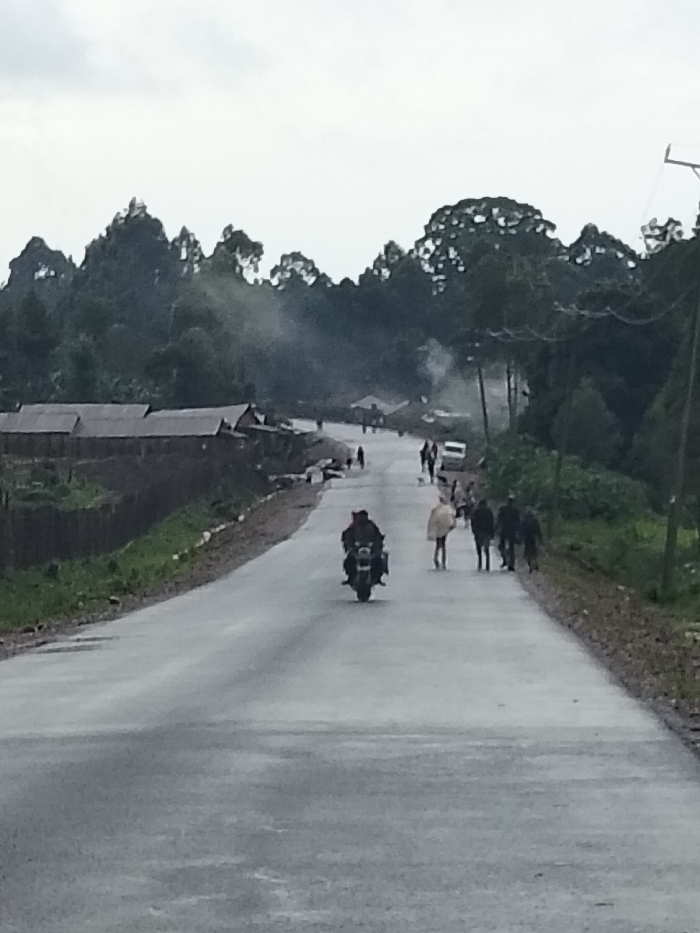 The scene depicts a public road in Nyansiongo ward, Kenya, under an overcast sky. A grey, possibly wet, paved road extends diagonally from the foreground to the midground. A dark-colored motorcycle, viewed from the rear, travels away from the observer in the center of the road. Ahead of the motorcycle, a group of approximately eight individuals are visible walking or standing on the road and its immediate shoulders. One person in the group wears a light-colored garment. Further down the road, a white vehicle, possibly a van or car, is partially visible on the right side. To the left of the road, a series of simple structures with corrugated metal roofs are visible, backed by dense green vegetation. To the right, a grassy shoulder transitions into a dense forest line. Plumes of light grey smoke rise from multiple points within the background foliage and structures, indicative of burning activity. A utility pole with overhead wires is present on the far right.