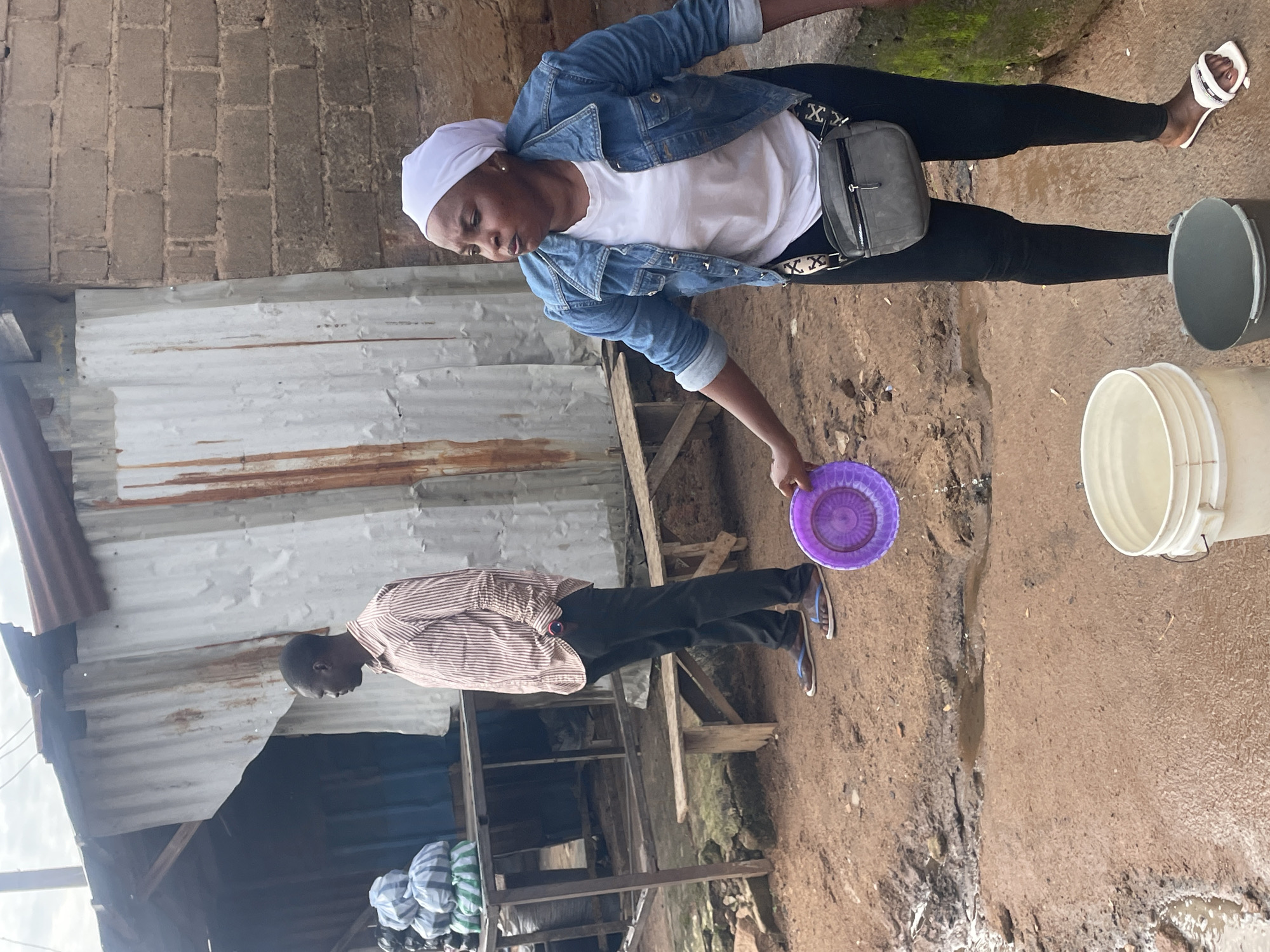 In Jos, Nigeria, a woman wearing a white headscarf, a blue denim jacket over a white t-shirt, black leggings, and white sandals, is holding a purple plate from which water is spilling. In front of her are two buckets, one white and one gray, on a muddy ground. Behind her, a man in a striped shirt and black trousers stands with his hands in his pockets, near a structure made of corrugated metal and brick. Bags of goods are visible on a table to the left. The scene depicts an outdoor setting with a dirt ground, indicating a potentially rural or underdeveloped urban environment.