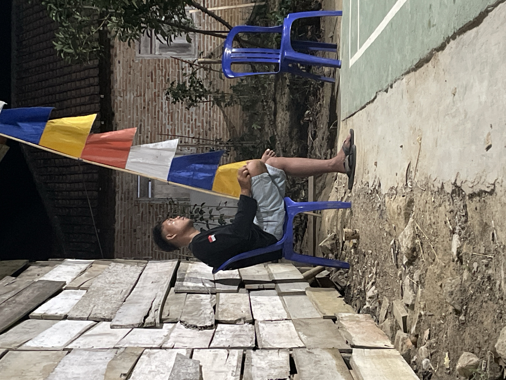 A young man is seated on a blue plastic chair on a concrete surface next to a dirt area. He is facing to his right, looking away from the camera. He is wearing a black long-sleeved shirt and light blue shorts. A flag pole with multiple colored flags (blue, yellow, red, white, and blue) stands to his right. Behind him is a wall constructed of horizontal wooden planks. Further in the background, there is a brick building with a tiled roof, a tree, and another blue plastic chair. The scene appears to be outdoors, likely in a residential or communal area in Kagungan Ratu, Indonesia. The lighting suggests it is evening or night.