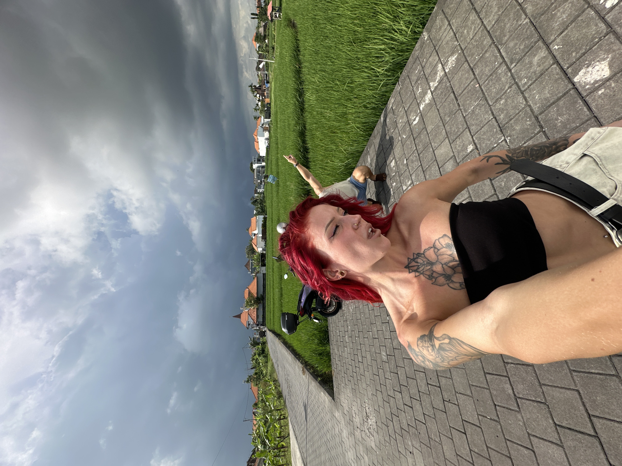 A waist-up photograph, rotated counterclockwise, shows a woman with red hair and numerous tattoos, wearing a black top and denim shorts with a black belt. She is holding the camera at arm's length, taking a selfie. In the background, a man sits on the edge of a rice field with his arm raised. The scene takes place on a paved walkway. In the distance, there are buildings with terracotta roofs, consistent with the architecture of North Kuta, Indonesia. A purple scooter is parked on the walkway behind the woman. The sky is overcast with dark clouds. Rice plants are visible along the walkway.