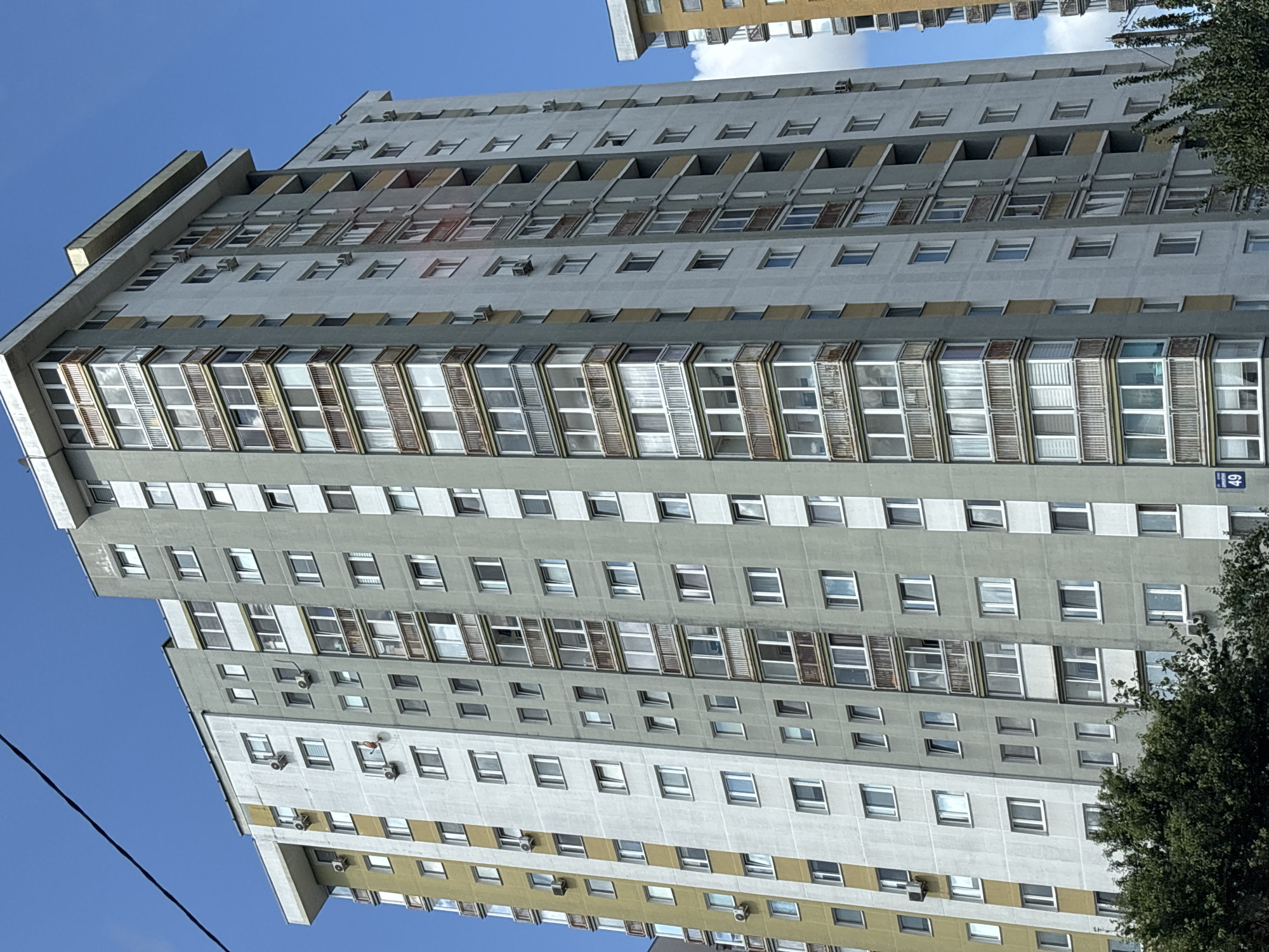 The image shows a high-rise apartment building in the city of Minsk, Belarus, under a bright blue sky. The building is tall and appears to be of Soviet-era architecture, with a mix of grey and yellow facade panels. Numerous windows and balconies, some enclosed with glass and others with wooden railings, are visible across the building's many floors. Several outdoor air conditioning units are mounted on the exterior walls. The bottom of the building is partially obscured by lush green trees. Another similar apartment building can be seen to the right, also under the clear sky. The overall scene depicts a typical residential area in Minsk.