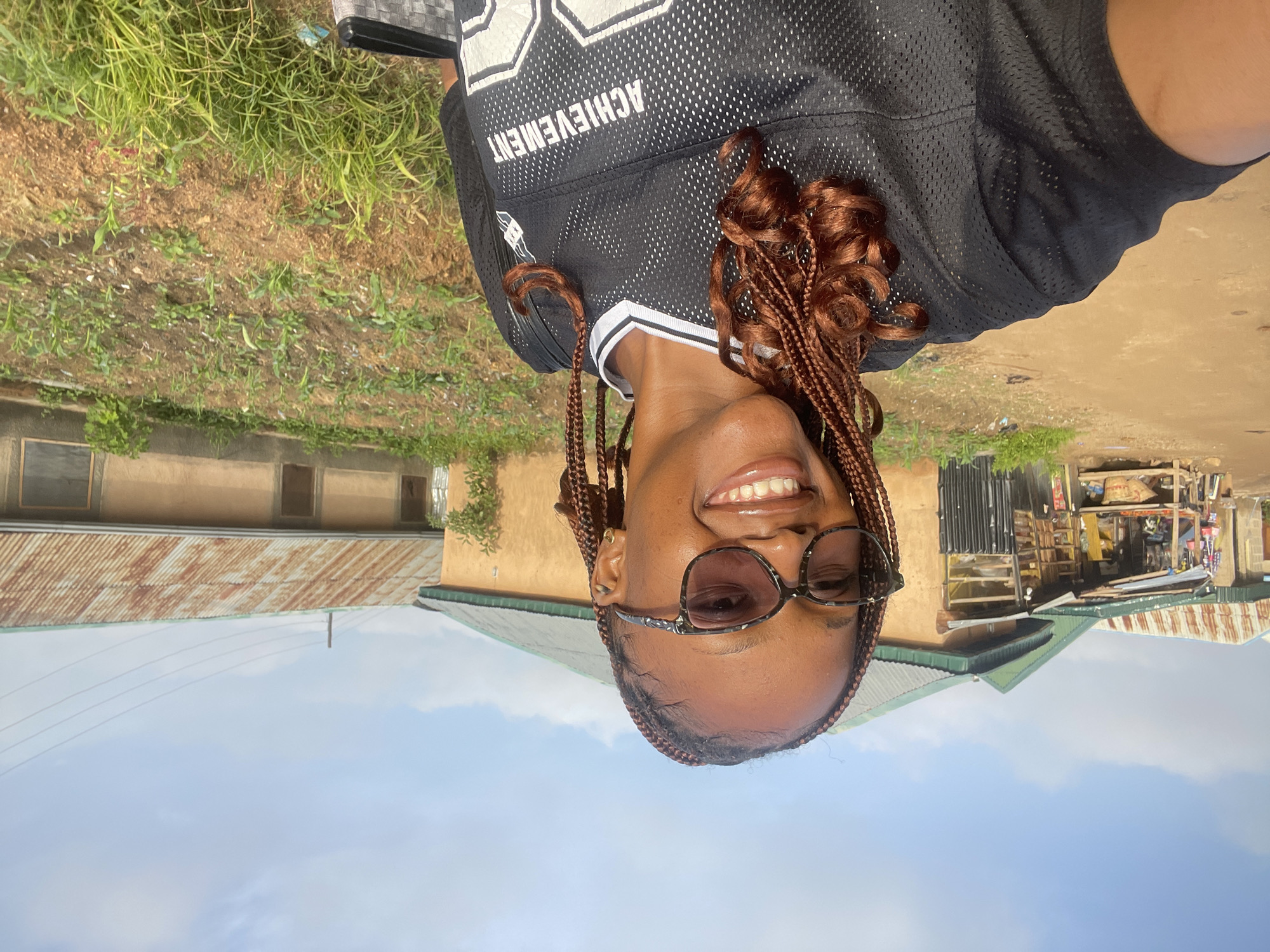 A young Black woman with braided auburn hair and tortoiseshell glasses smiles at the camera. She is wearing a black jersey with white trim and the word "ACHIEVEMENT" printed in white across the chest. She appears to be standing in a dirt yard in front of a row of buildings with corrugated metal roofs. The buildings seem to be small shops or residences, one with goods displayed outside on shelves. A patch of green plants, possibly a small garden, is visible to the right of the woman. The sky is blue with some scattered clouds. The overall impression is a casual, outdoor scene in a village or rural area.