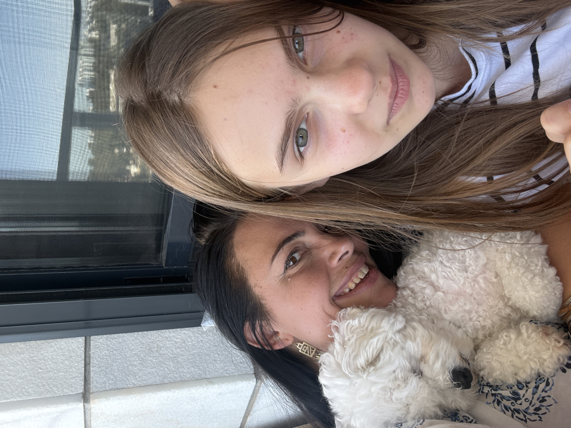 The image shows a selfie of two people and a small white dog. The person in the foreground is a woman with dark hair, smiling and looking to her right. She is wearing a patterned top. Next to her, and slightly behind, is a young girl with long, light brown hair. She is also smiling and looking towards the camera. She is wearing a striped shirt. Curled up between the two people is a fluffy white dog. The background appears to be outdoors, possibly on a balcony or patio, with a window frame and textured wall visible. The lighting suggests it is daytime. The overall mood of the image is happy and relaxed, with both individuals and the dog appearing content. There is no visible text in the image.