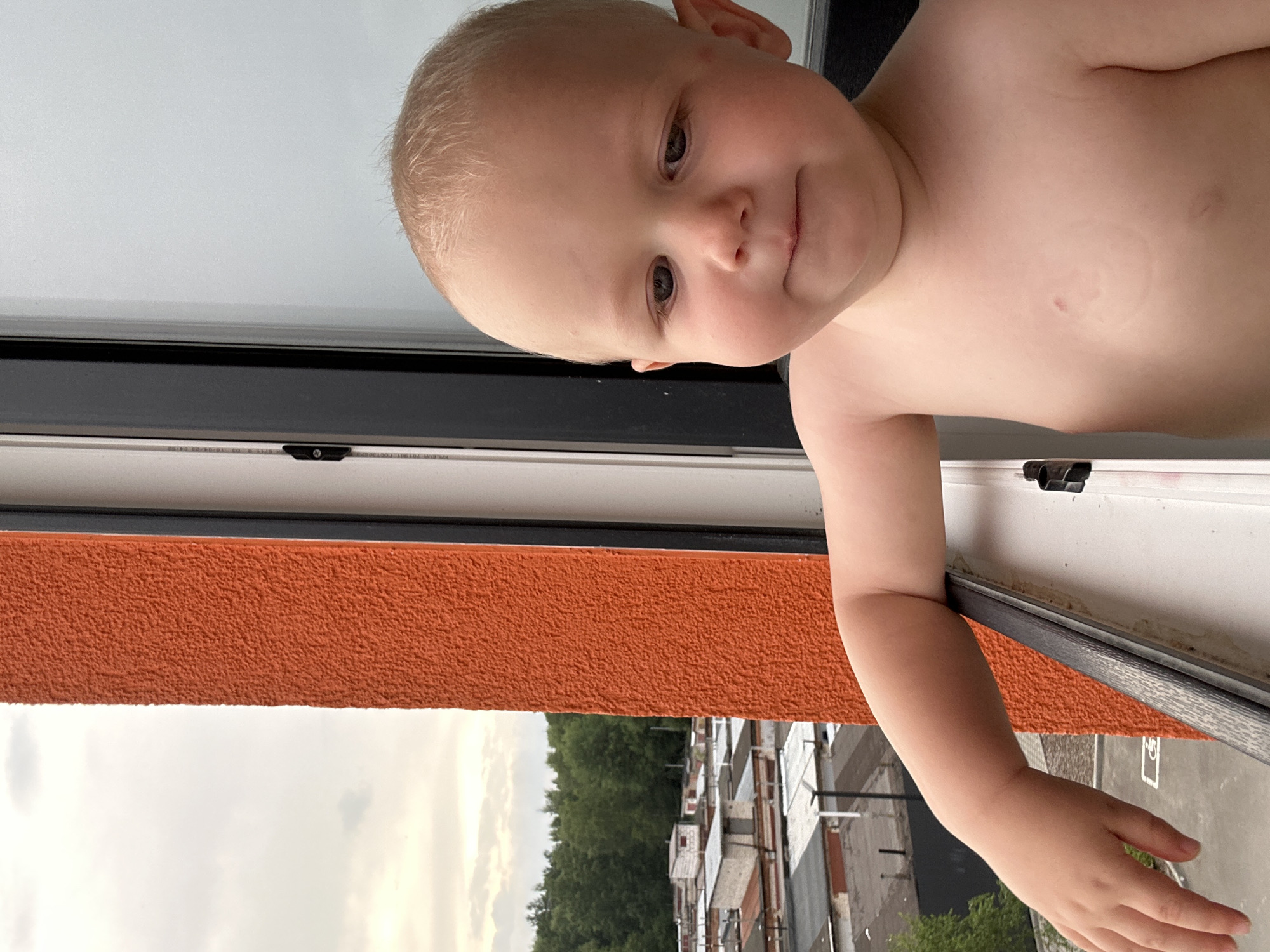 A fair-skinned child with blond hair and blue eyes is pictured from the chest up, looking down and to the right. The child is partially leaning out of a window, with their right arm extended downwards and their left shoulder visible. The background to the left of the child shows an orange textured wall and beyond that, a view of a city or town with buildings and trees. The sky is overcast. The image appears to be taken outdoors, possibly on a balcony or from a window of an apartment building. The child's body is bare from the chest up. The child's skin is smooth, with some redness on the chest. The background view is slightly out of focus. The child's expression is neutral, with their gaze directed downwards. The overall lighting suggests it is daytime, but the overcast sky creates a diffused light. The context of Kaluga, Russia is not explicitly conveyed by visual elements in the image itself but is provided as external information.