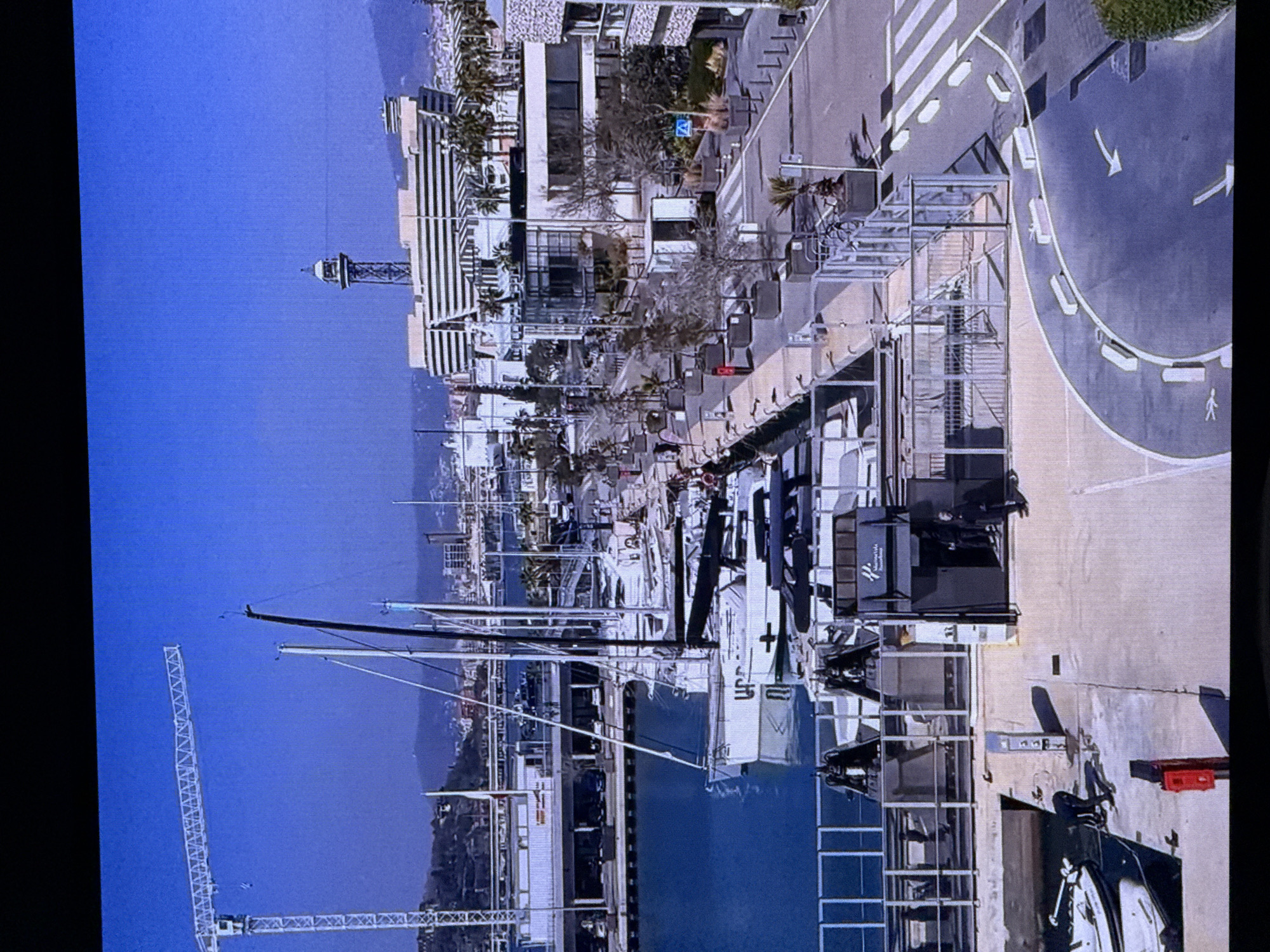 This media file captures a view of a harbor with several sailboats docked, alongside modern buildings and a prominent tower. In the foreground, a large white construction crane stands tall against a clear blue sky. The scene is a vibrant harbor, likely a marina, characterized by the presence of yachts and the surrounding urban development. The time of day appears to be daytime, with bright sunlight casting shadows, suggesting a clear and sunny weather condition.
The modern architecture of the buildings, including a striped facade and glass windows, indicates a contemporary urban setting. A tall, slender tower with a distinctive observation deck is visible in the background. The harbor itself is filled with the masts of sailboats, suggesting they are either moored or undergoing maintenance. Additional structures like walkways, railings, and what appear to be parts of a building complex with metal frameworks are also present.
In the background, a distant cityscape and hills or mountains can be seen, hinting at a coastal location. The overall impression is one of a bustling port and a developed urban area.
The visible text in the image includes a small blue sign with a white symbol on a pole, and some text on a black box structure, which is partially obscured. The location is identified as Dubai, United Arab Emirates. No specific emotions are conveyed, and the activity appears to be the normal operation of a harbor and its surrounding urban environment.