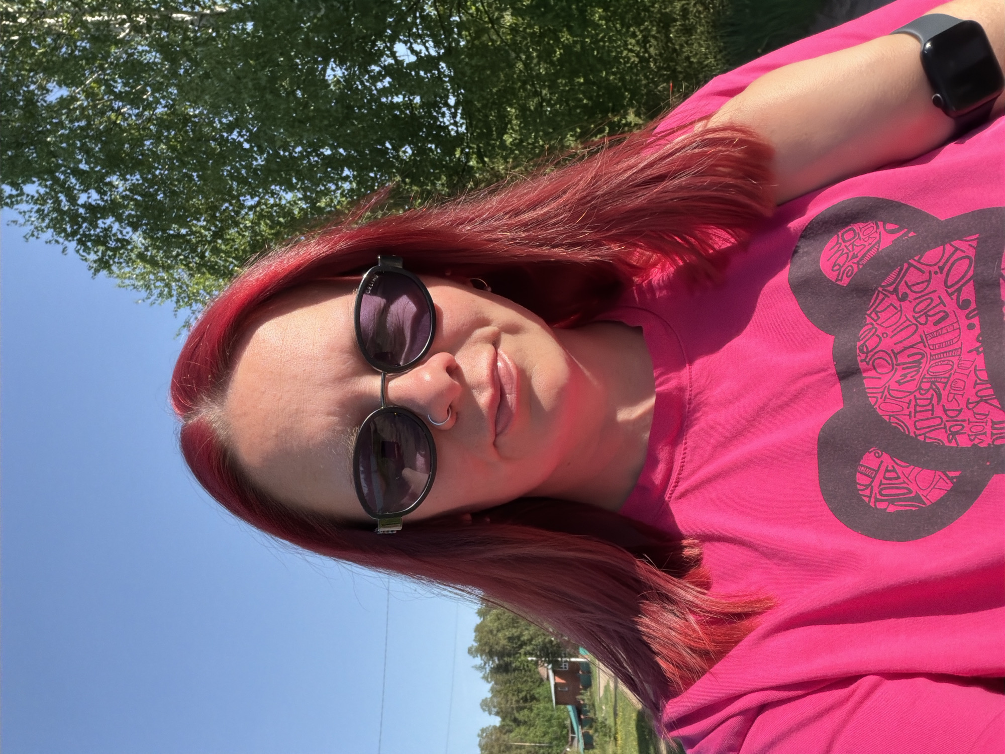A woman with red hair, sunglasses, and a nose ring is wearing a pink t-shirt with a dark gray graphic. She has a smartwatch on her left wrist. The background features a blue sky, trees, and a glimpse of a house. The photo appears to be taken outdoors in a sunny environment, potentially in the vicinity of Chaikovsky, Russia, based on visual cues suggesting a residential or semi-rural area.