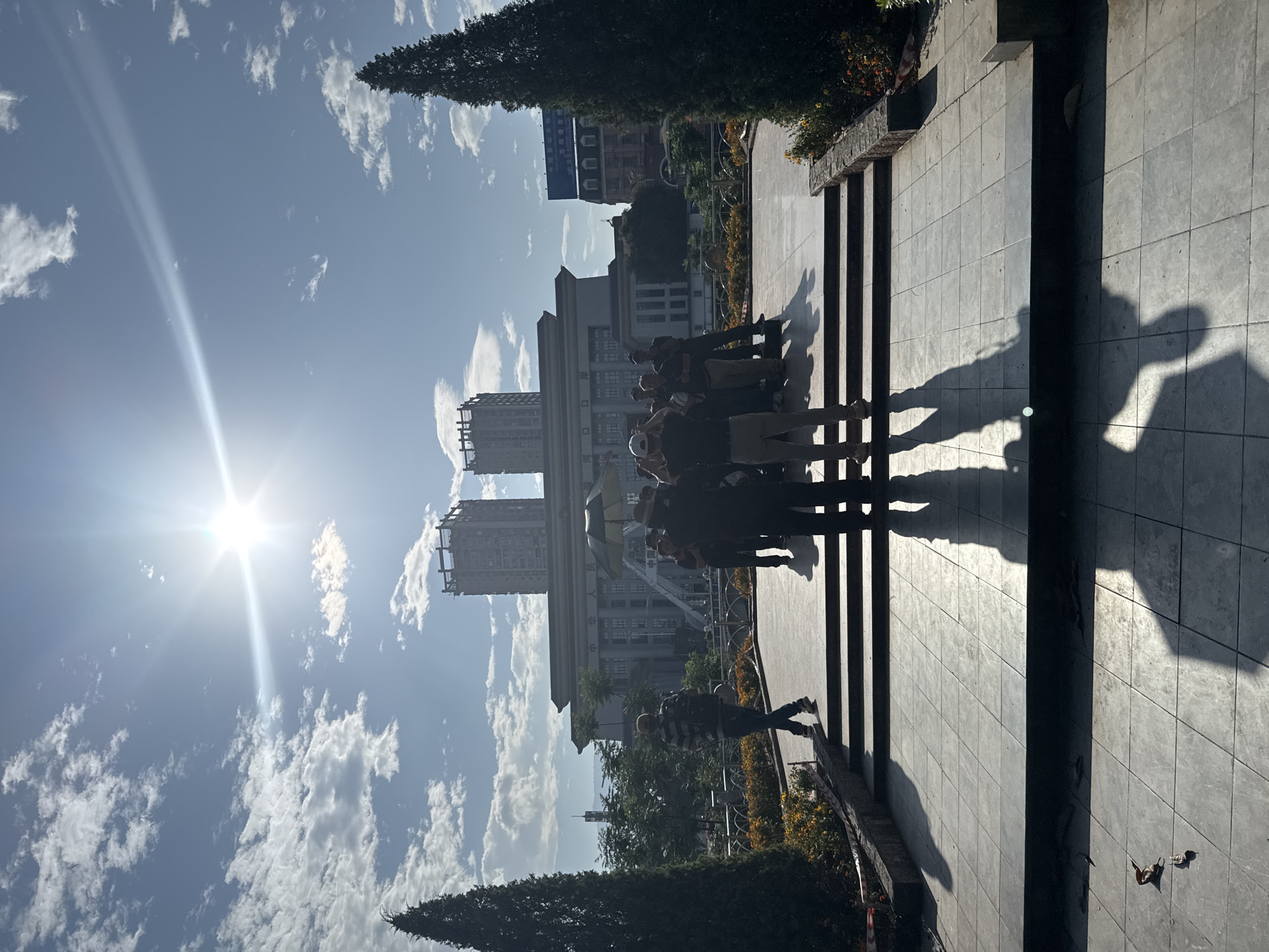 The image shows a group of people standing on a set of stairs in Lao Cai Ward, Vietnam, with a large building complex in the background under a bright sky with scattered clouds. The sun is shining brightly. There are trees and flowers visible near the base of the stairs. A person is walking along the edge of the stairs. Shadows of people are cast across the paved area. A tall building is also present behind the large building.