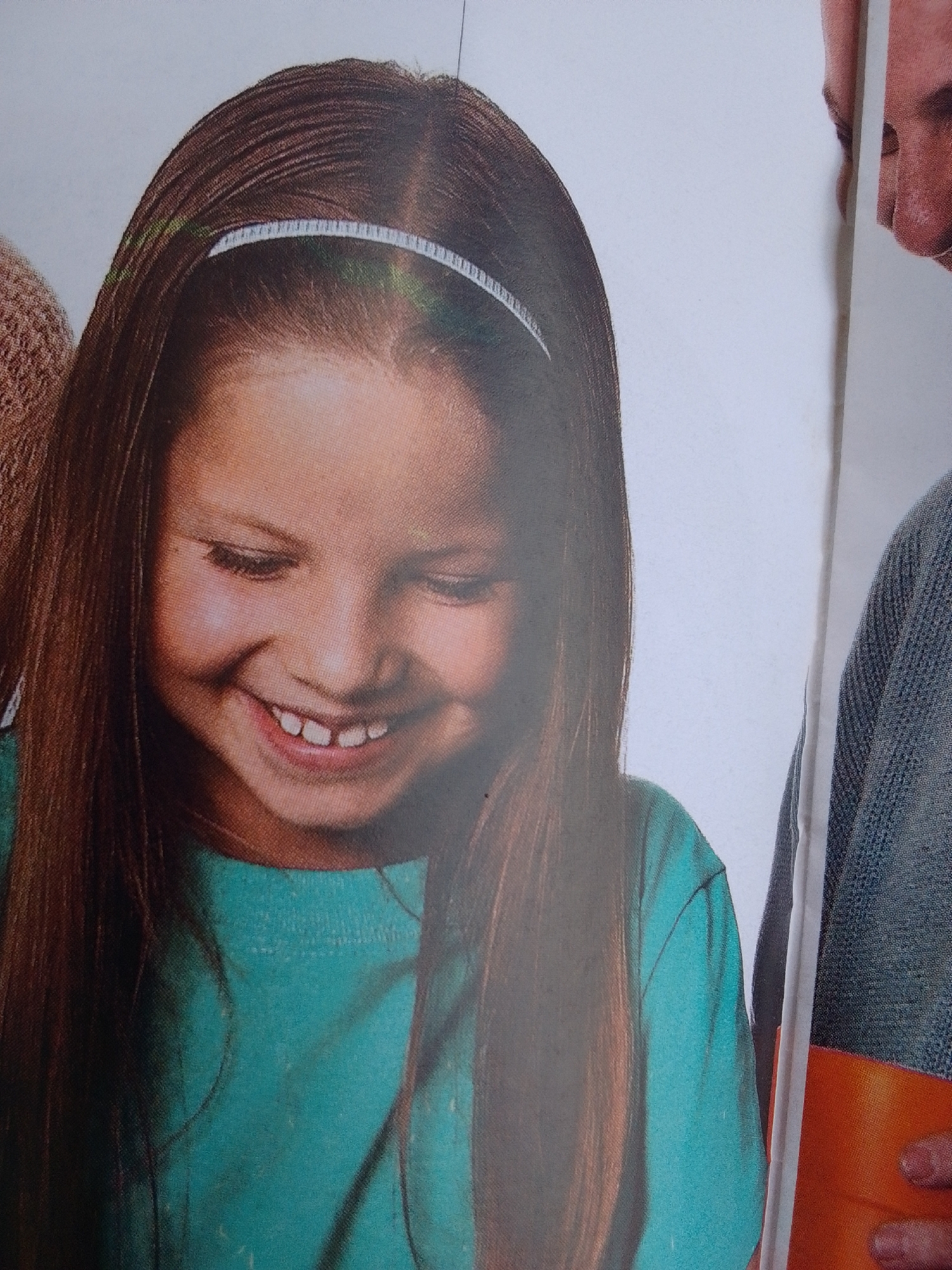 This is a close-up photograph of a young girl with long brown hair and a teal t-shirt. She is wearing a white headband. She is smiling with her mouth open, showing her teeth. Her eyes are closed. The background is a plain white surface. To the right of the girl, a portion of an adult's face and shoulder is visible. The adult appears to be wearing a gray sweater. There is also a bright orange object visible in the lower right corner. The image appears to be a candid shot, possibly from a family gathering or a photoshoot. The lighting is bright and even, suggesting an indoor setting or an overcast day. The overall mood is cheerful and happy. There is no visible text in the image.