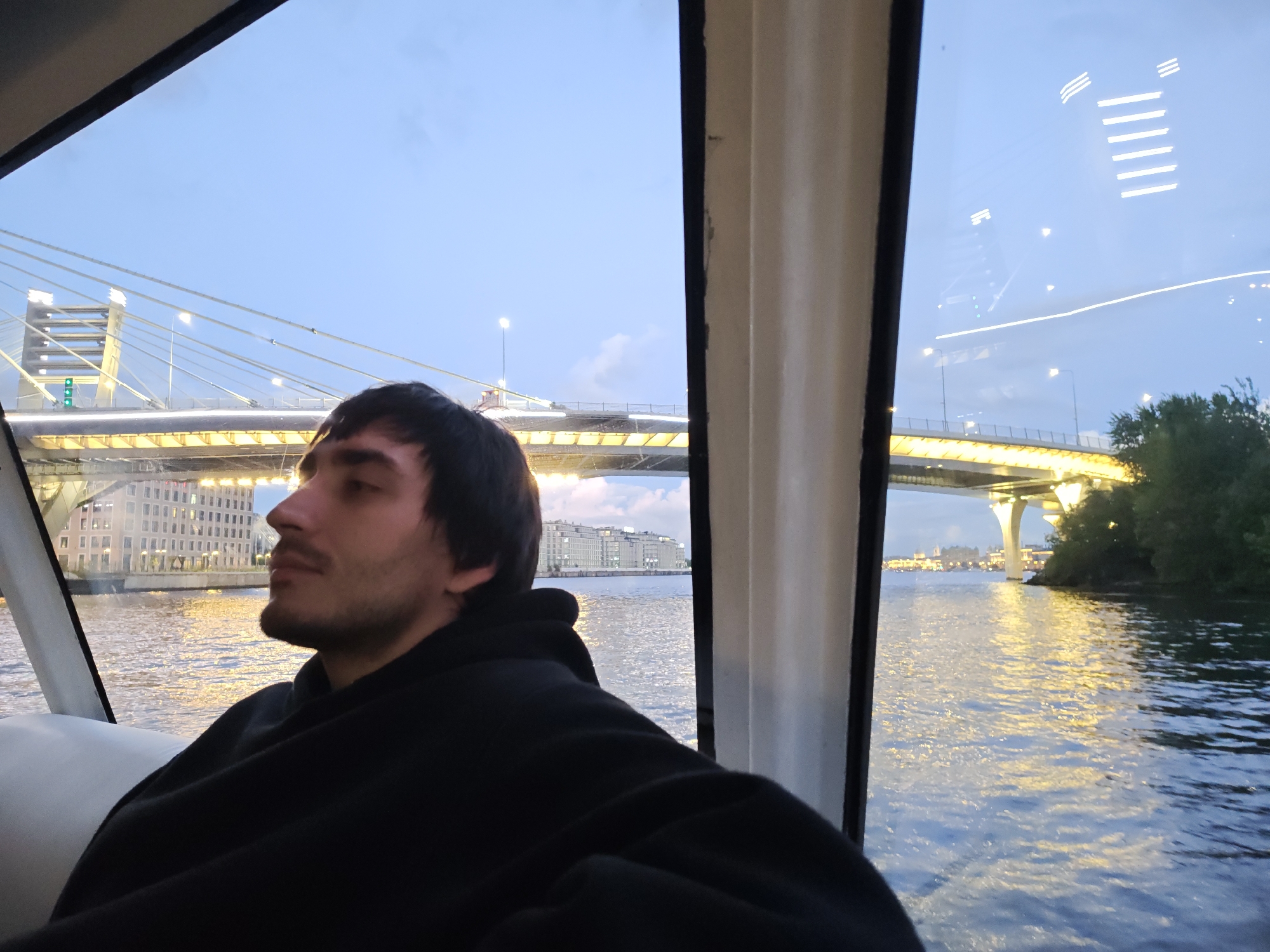 An outdoor, wide shot captures a man in a black hoodie seated on a boat at dusk, looking out over a river towards a city landscape. The man's profile is visible, with dark hair and a beard. In the background, a large illuminated bridge spans the river, with buildings lining the far bank and trees on the right. The water reflects the lights of the city and the bridge, creating a shimmering effect. The sky is a deep blue, indicating the transition from day to night. The overall scene suggests a serene evening boat ride in Saint Petersburg, Russia, likely along the Neva River, given the architectural style of the bridge and buildings which are characteristic of the city.