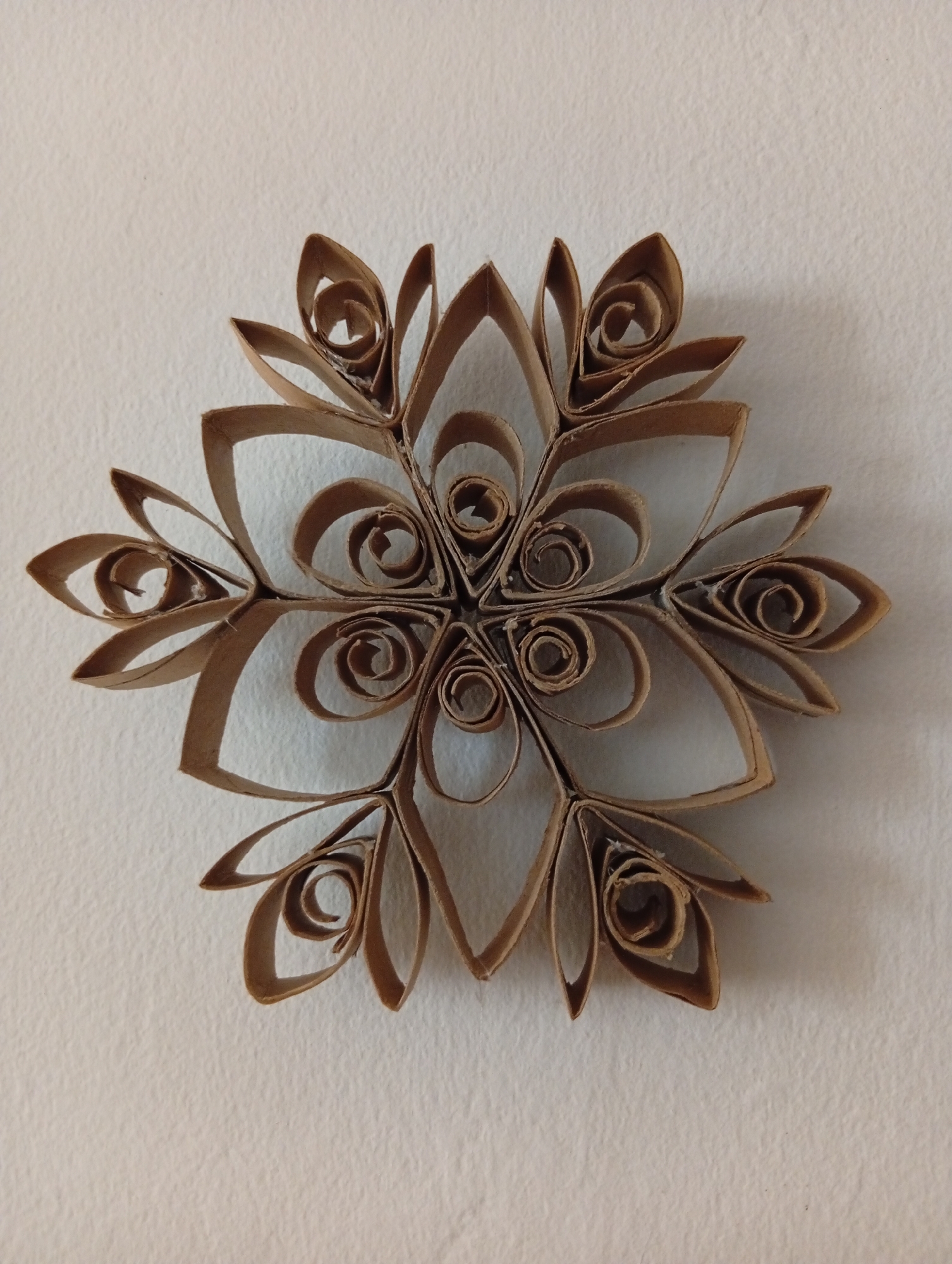 A brown, star-shaped craft made of rolled cardboard is affixed to a textured, off-white wall. The craft is symmetrical, with six points, each featuring multiple layers of curled cardboard creating a layered, petal-like effect. The center of the craft has smaller, tightly wound curls. The lighting suggests the object is illuminated from above, casting subtle shadows on the wall. There are no people or other objects visible. This image was taken in San Antonio de Los Altos, Venezuela.