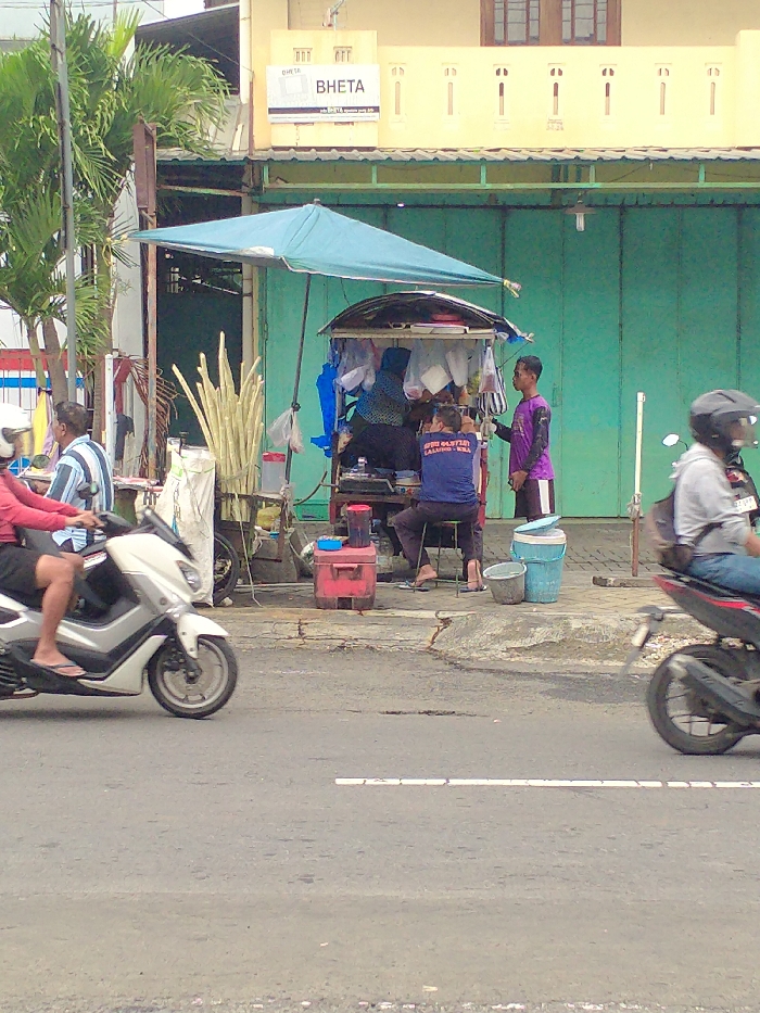 This daytime image captures a vibrant street scene in Karanganyar, Indonesia, showcasing a bustling roadside sugarcane juice stall. The main subject is a vendor's cart, shaded by a prominent blue umbrella, with numerous long sugarcane stalks piled high, ready for processing.
At least five individuals are visible: two people on scooters are in motion on the asphalt road in the foreground—one in a red shirt on a white scooter, and another in a grey hoodie on a dark scooter. Near the sugarcane stall, a person with a head covering appears to be working behind the counter. Next to them, a man in a blue t-shirt sits on a stool, possibly a vendor or patron, with his shirt displaying the text "SPESIAL ES TEBU LALUNG - KRA," indicating "Special Sugarcane Ice" and a specific location (Lalung, Karanganyar). Another man in a purple shirt stands nearby, looking towards the stall. Further left, another man in a striped shirt stands beside a white cart that also holds sugarcane stalks.
The setting includes a building in the background with a light yellow upper facade and closed green metal doors on the lower level. A sign on this building clearly reads "BHETA," with additional smaller text "info BHETA" and "bhetaproduction only 010." Green palm trees are visible on the left side, adding a tropical element to the urban environment. The bright lighting suggests it is a clear, sunny day. The overall scene depicts typical daily life and street commerce in the region.