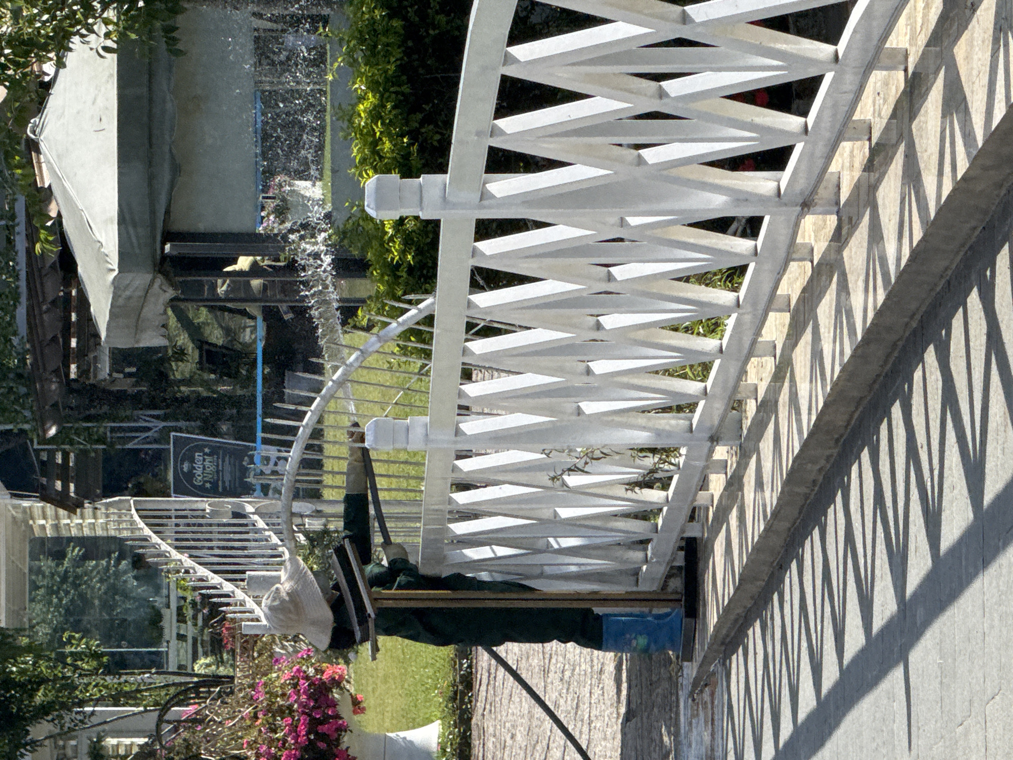 In Da Lat, Vietnam, a person wearing a straw hat and a green uniform is cleaning a white railing on a bridge. The person is also wearing blue boots and gloves, and is using a cleaning tool that appears to be a squeegee on a long handle. Water is spraying from a hose in the background, and there are pink flowers and green foliage surrounding the area. The sunlight casts sharp shadows on the bridge's surface, creating a geometric pattern. A sign with "Golden Night" written on it is visible in the background, along with a white tent-like structure and trees.