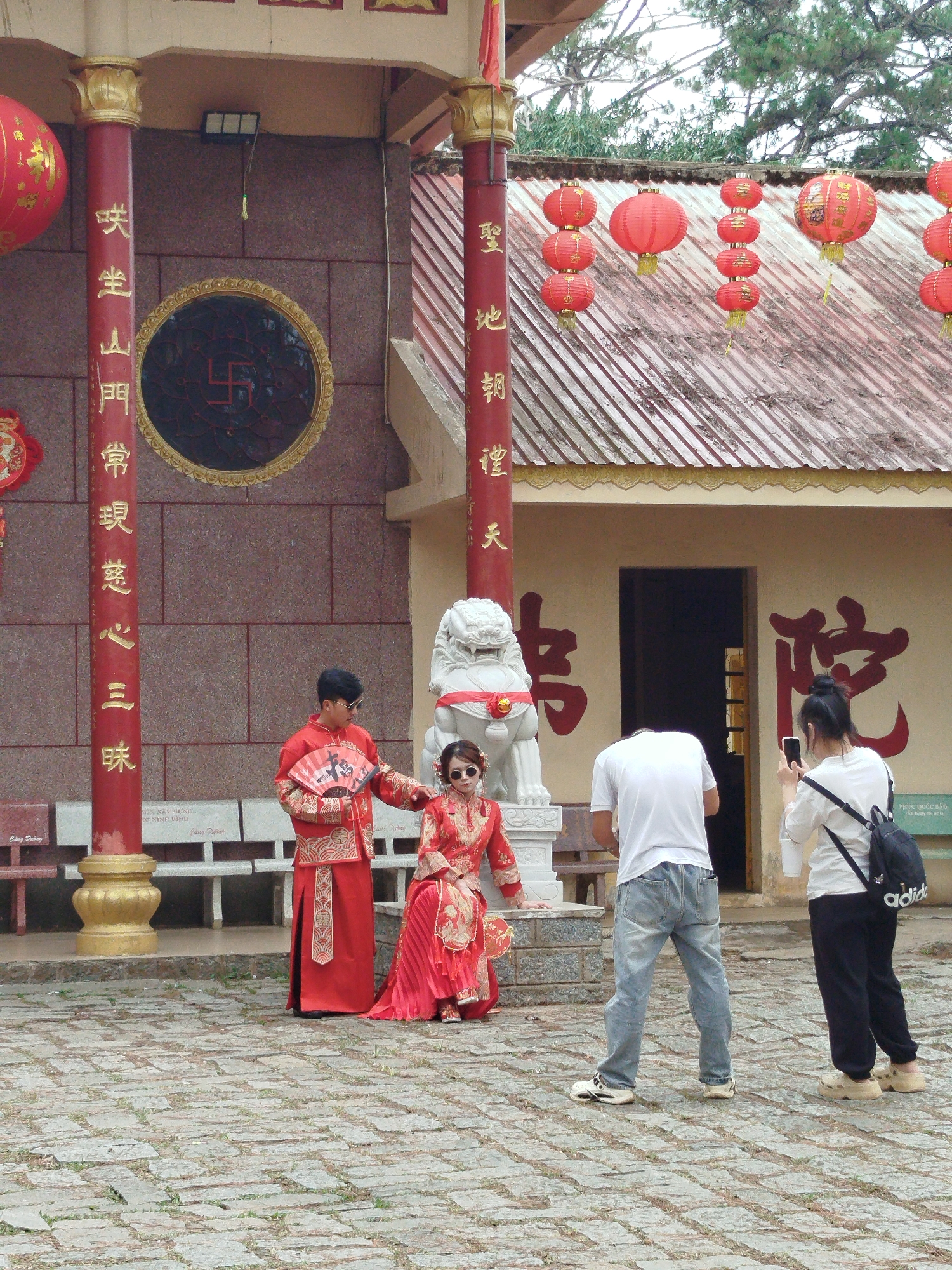 The image depicts a couple dressed in traditional red Vietnamese clothing posing for a photo in front of a Buddhist temple. The temple has red columns with gold lettering, a large window with a swastika symbol, and red lanterns hanging from the roof. A white stone lion statue sits near the entrance. Two people are taking photos of the couple with their phones. The scene is likely taking place in Da Lat, Vietnam, given the temple's architecture and the couple's traditional attire, which is often worn for weddings or other special occasions in Vietnam.