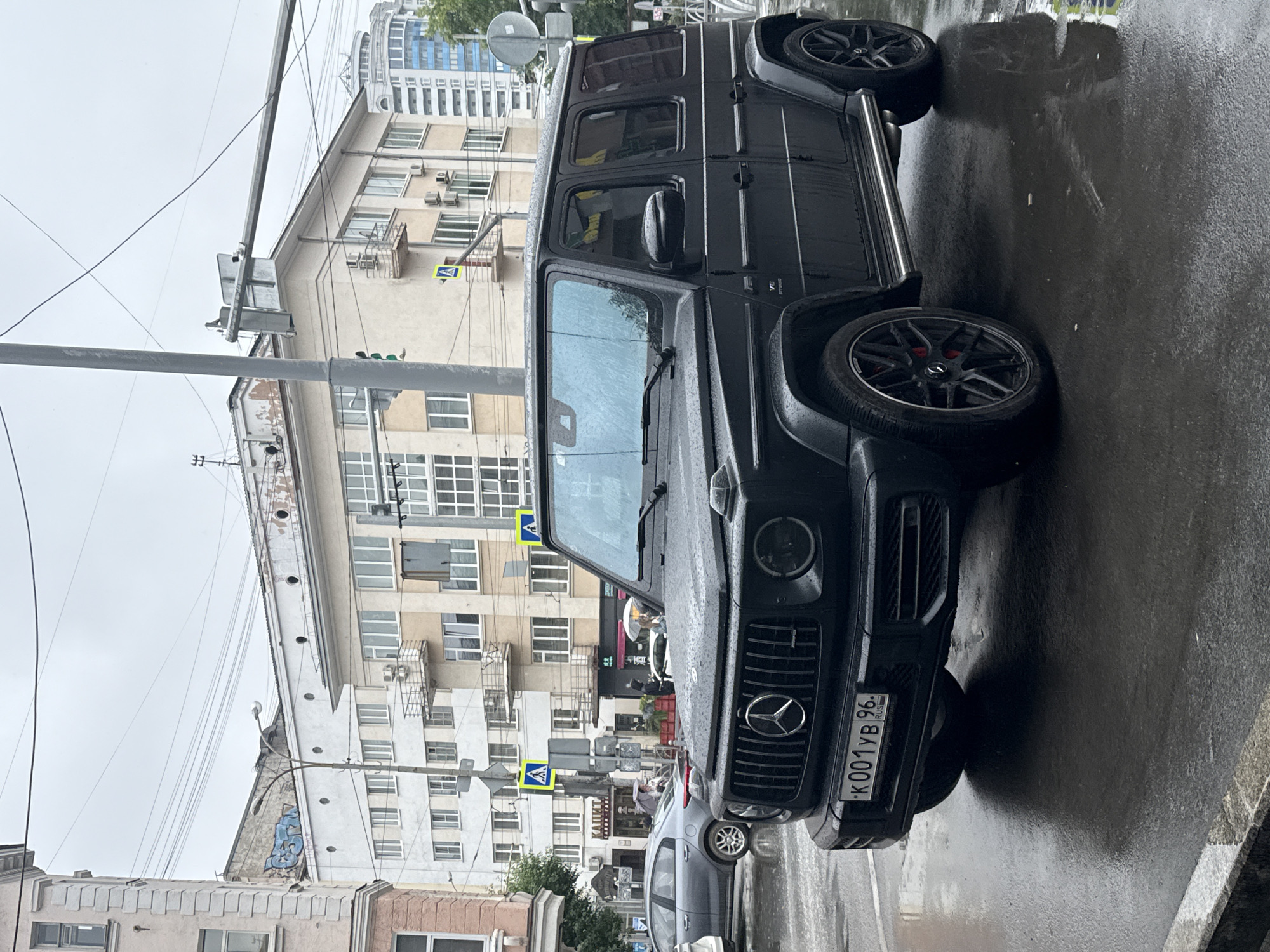 A black Mercedes-Benz G-Class is parked on a wet, overcast street, likely in Yekaterinburg, Russia. The vehicle's tires are visibly wet, and the surrounding pavement reflects the dull, cloudy sky, indicating recent or ongoing rain.
The G-Class, a prominent object in the foreground, has its license plate visible: "K001YB 96" with "RUS" below it. The car's grille features the Mercedes-Benz star logo. The vehicle is angled towards the viewer, with its side and front prominently displayed.
In the background, several multi-story buildings line the street. Some have light-colored facades and numerous windows. One building appears to have graffiti in blue on its side. Traffic signs are visible, including yellow pedestrian crossing signs. Power lines crisscross the sky.
A few other vehicles are present, including a silver car parked behind the G-Class. A person, partially obscured, can be seen near the silver car. Another figure is visible on a sidewalk in the distance, under an umbrella. The overall atmosphere is somber and damp, consistent with a rainy day in an urban environment. The time of day is not explicitly discernible but appears to be daytime due to the ambient light.