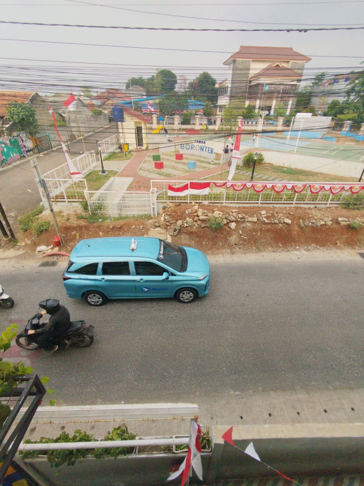 The image shows a street scene in Depok, Indonesia. In the foreground, a blue Bluebird taxi is driving on the road, and a person on a motorcycle is also visible. In the background, there is a park with a basketball court, a building with a brown roof, and houses. Indonesian flags are displayed throughout the scene, suggesting a celebration or national event. The sky is overcast, and the overall atmosphere is one of urban activity.