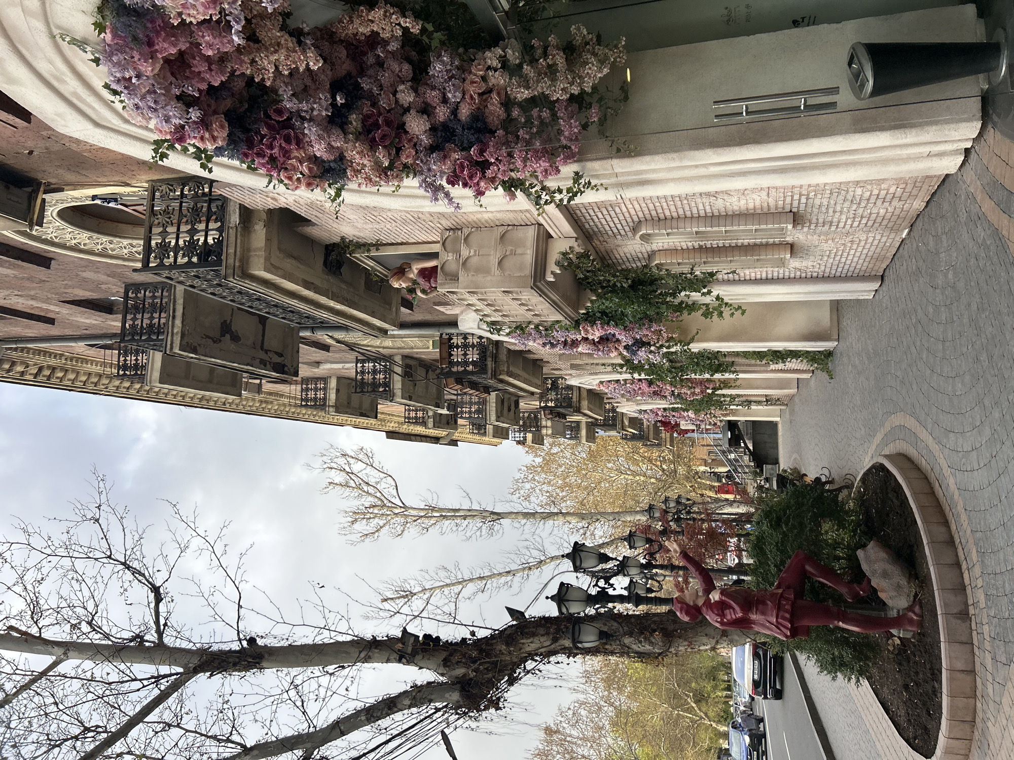 The image depicts a street scene in Yerevan, Armenia. The buildings lining the street have architectural details like balconies and ornate window fixtures, some with floral arrangements. A pedestrian walkway with patterned brickwork runs alongside the buildings. A sculpture of a figure in a red outfit lies within a circular planter. Overhead, bare tree branches stretch out against a cloudy sky, and street lamps are interspersed among the trees. Cars are parked along the street, and at least one person is visible on a balcony.