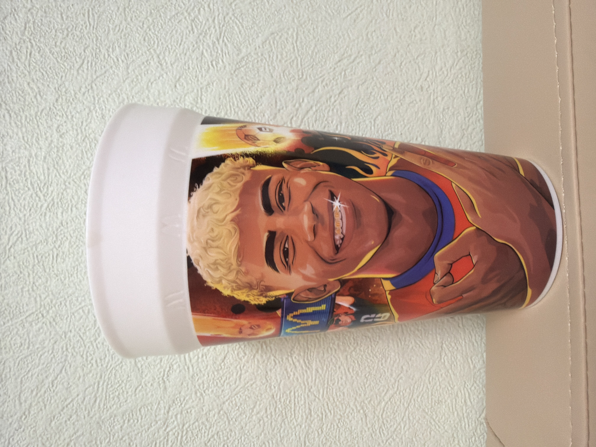 A white cup with a cartoonish image of a young man is depicted. The man has blonde curly hair, is smiling, and is wearing braces with diamonds. He is wearing an orange and blue jersey. The cup has a white rim. The background is a textured, light-colored wall. The cup is sitting on a light brown surface.