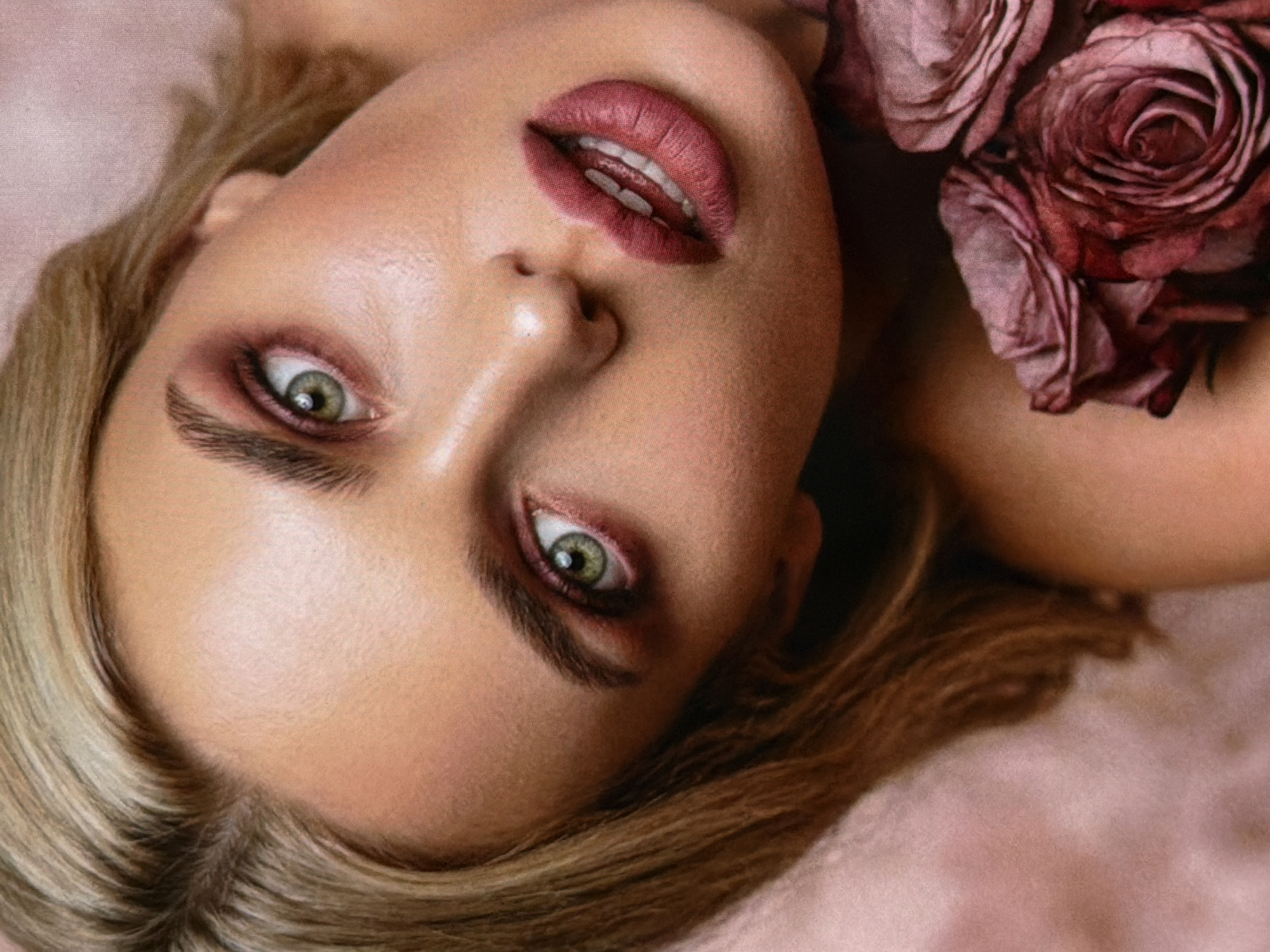The image is a close-up portrait of a woman with blonde hair and green eyes. She is wearing dark pink eyeshadow and lipstick. Her head is tilted back, and she is looking up. A bouquet of dark pink roses is held near her shoulder. The background is a soft, blurry pink. There is no indication of the city of Bobrovo, Russia, in this image.