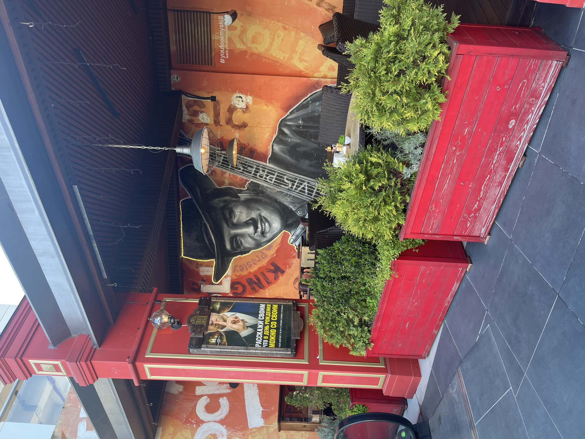 The image shows an outdoor seating area, likely a cafe or restaurant, possibly in Sochi, Russia, based on the Cyrillic script. The scene is dominated by a vibrant orange wall featuring a large mural of Elvis Presley with a guitar. Adjacent to the mural is a red, vintage-style wall that contains an advertisement with Cyrillic text.
To the right of the wall are three large, red rectangular planters filled with green shrubbery. The planters sit on dark-colored, tiled flooring. The seating area appears to be covered, as evidenced by the structural beams overhead. There is an outdoor lamp attached to the red wall. The scene appears to be a mix of vintage and modern elements, creating a unique aesthetic.