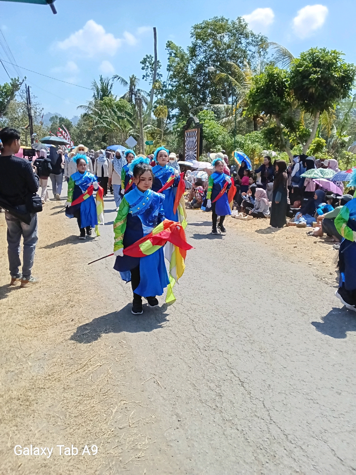 On a sunny day in Ngadirenggo, Indonesia, a parade is in progress along a paved road lined with trees and vegetation. The central focus is a group of young girls in matching blue and green uniforms, adorned with colorful sashes and headpieces. They are marching in single file, each holding a baton with a flowing red and yellow flag attached. The girls appear to be performing, with their outfits and props suggesting a synchronized routine.
On either side of the road, spectators are gathered, many of them seated or standing behind barriers. They are a diverse mix of ages, with some individuals holding umbrellas for shade. The crowd comprises men, women, and children, some of whom are dressed in traditional Indonesian attire. Behind the spectators, more trees and foliage are visible, creating a lush, tropical backdrop. The sky is blue with scattered white clouds, indicating fair weather for the event. The overall scene depicts a community gathering, likely a cultural or celebratory event, marked by the vibrant parade and the presence of an engaged audience.