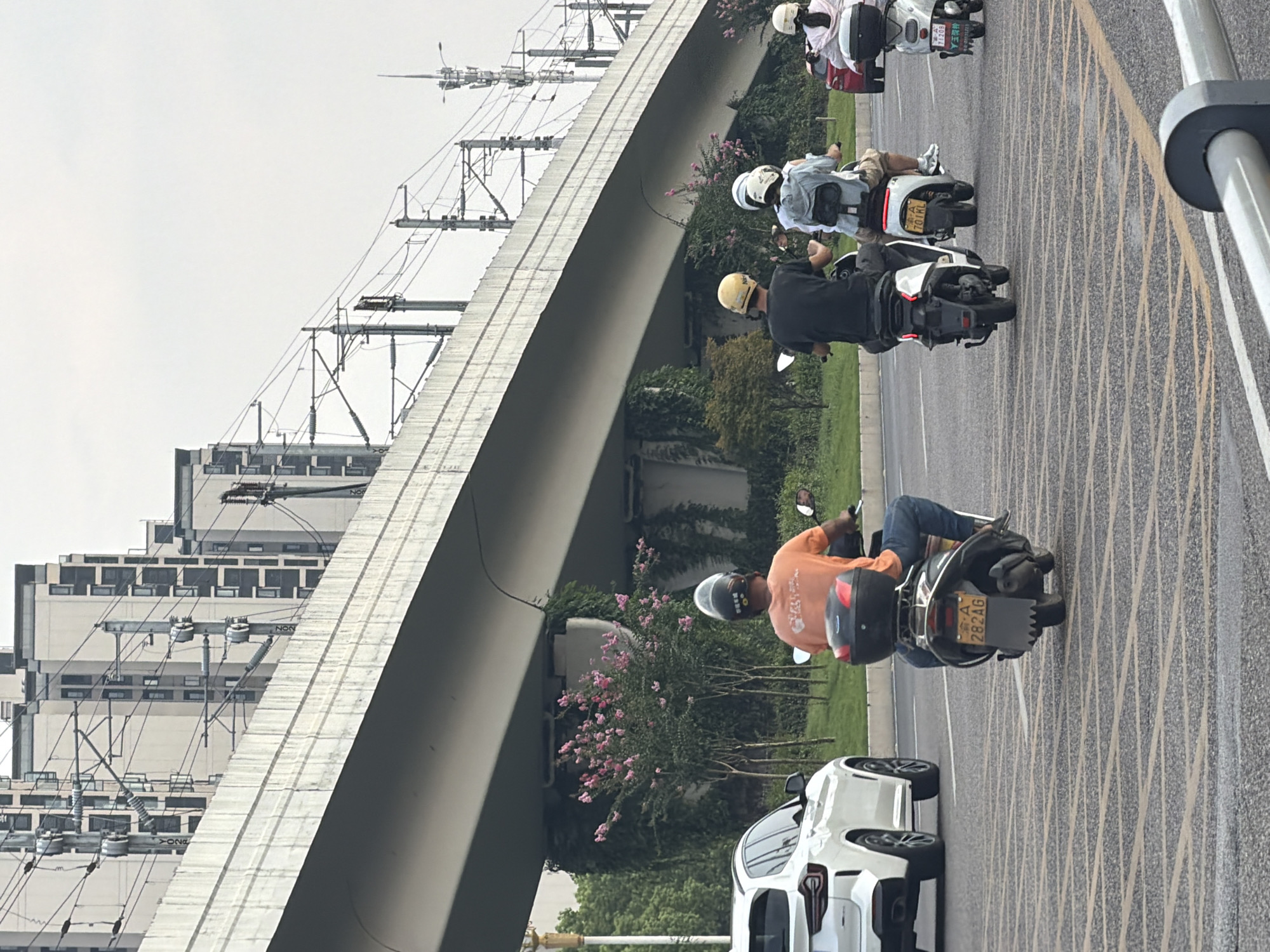 The image depicts a street scene in Jiangjin, China, featuring several individuals riding motorcycles and a white SUV. In the foreground, a person wearing an orange shirt and blue jeans rides a black scooter. Behind them, two individuals on a white scooter are visible, one wearing a yellow helmet and the other a light blue backpack. Further in the background, a person on a white scooter with a pink jacket is seen. A white SUV is parked on the left side of the road. The background includes a large elevated concrete structure, possibly a bridge or overpass, with electrical wires and utility poles running alongside it. Tall buildings are visible behind the elevated structure, and the environment appears to be a city street with greenery and flowering bushes under the overpass. The road surface is marked with yellow and grey lines.