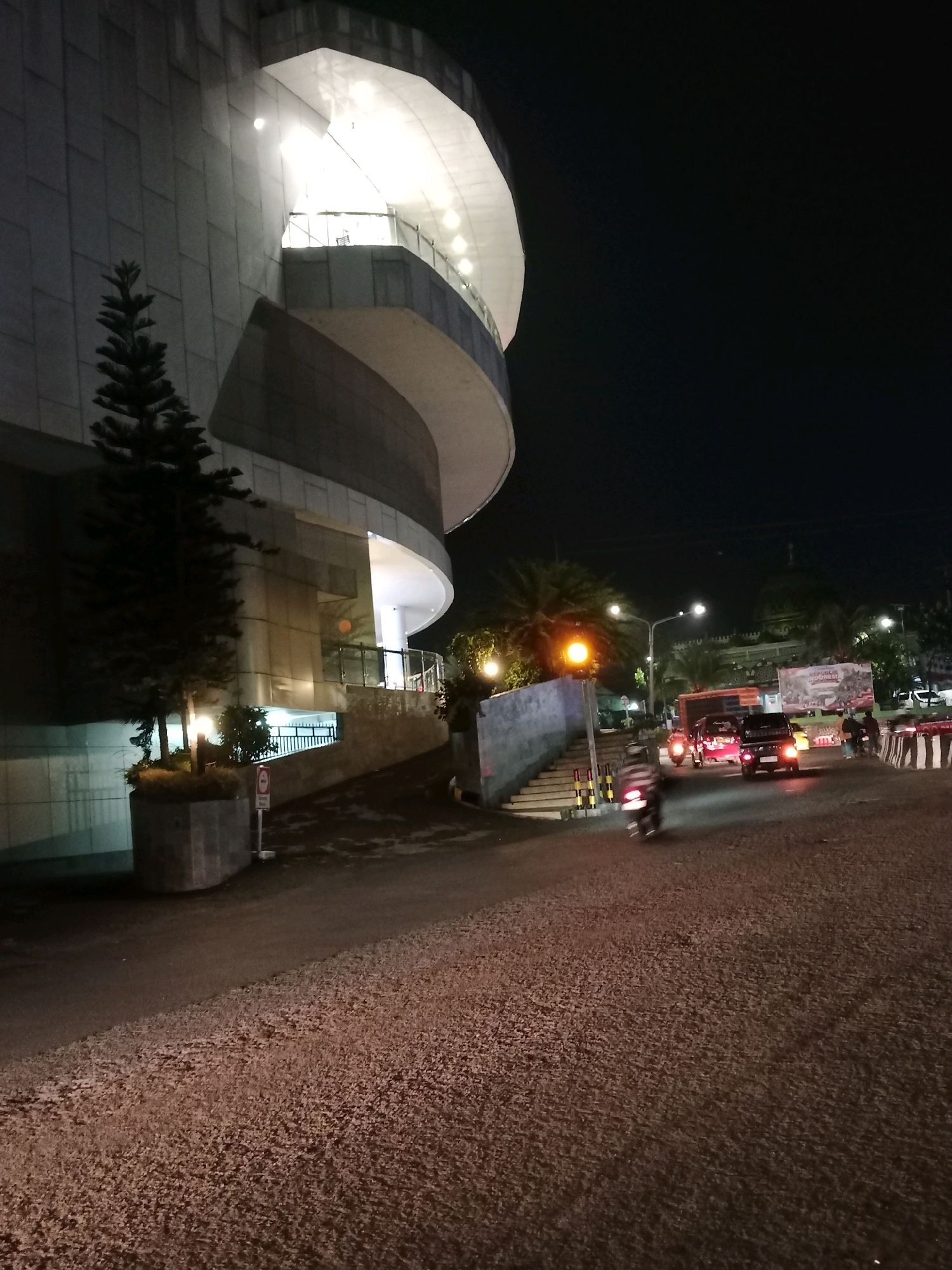 The image depicts a street scene at night in Cianjur, Indonesia, near a modern, multi-story building with illuminated floors. A lone evergreen tree stands in a planter in front of the building. The street is paved with asphalt and gravel, and there is a slight incline. Several vehicles, including cars and motorcycles, are visible on the road, with their headlights and taillights illuminated. Pedestrians are also present, walking on the sidewalk and crossing the street. A traffic light with a yellow signal is visible, as are various signs and barriers. The sky is dark, indicating nighttime.