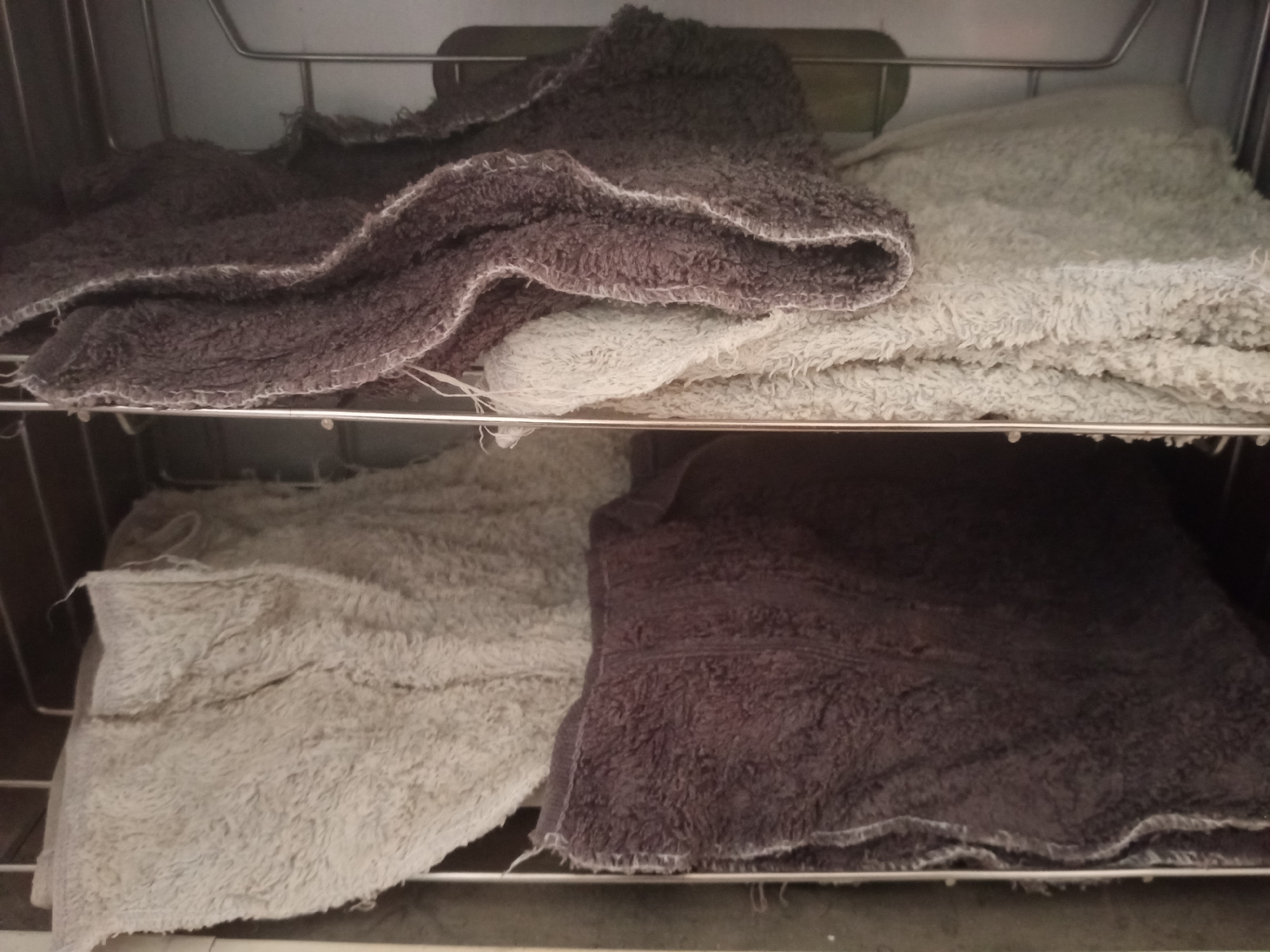 The image shows two shelves inside a metal rack. On the top shelf, a dark grey towel is folded over a light grey towel. On the bottom shelf, a light grey towel is folded on the left, and a dark grey towel is folded on the right. The towels appear to be plush and textured. The rack is metallic and appears to be in a storage area or a laundry room. The location of this scene in Jalingo, Nigeria is not provided in the image or its description.