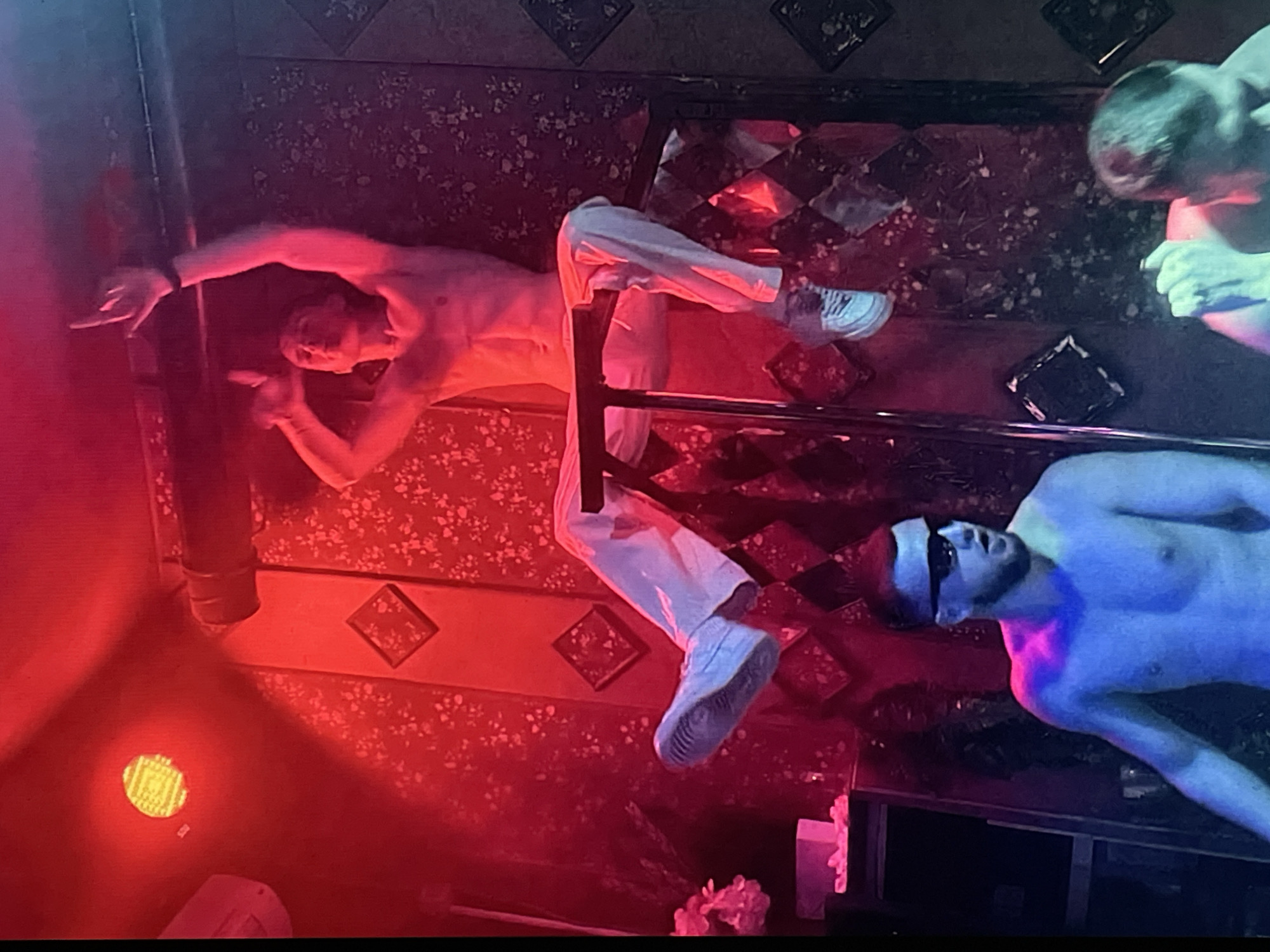 The image shows an interior space, possibly a nightclub or bar, in Saint Petersburg, Russia. Three shirtless men are visible. One man is reclining on a chair or ledge, wearing white pants and shoes. Another man is wearing dark sunglasses and is only partially visible. The third man is only partially visible in the upper right corner. The scene is illuminated by red and blue lighting. The background features decorative patterns on the walls and floor. A spotlight is visible on the floor in the lower left corner. There are also white floral decorations near the bottom of the image.
