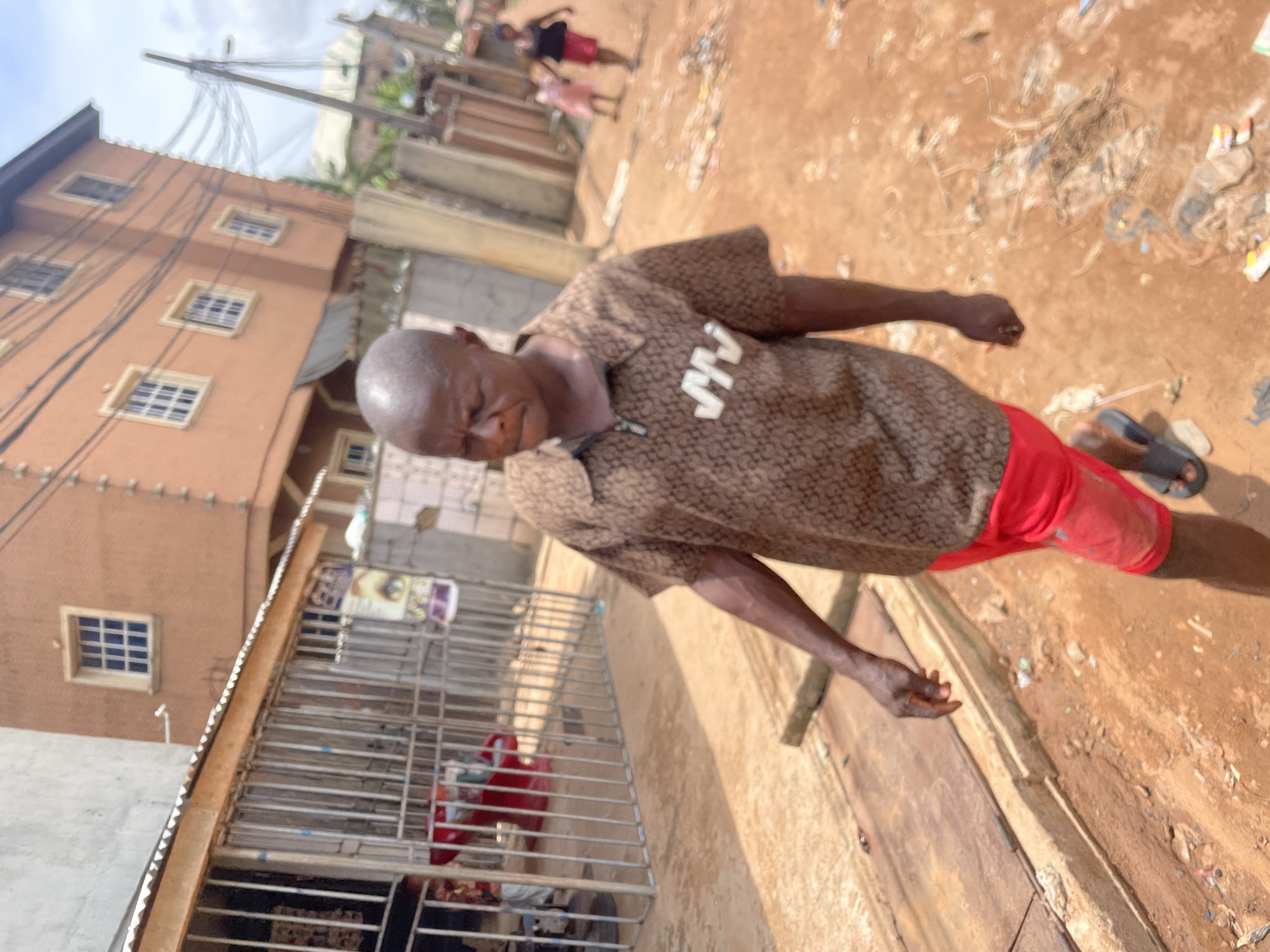 An elderly bald Black man wearing a patterned brown shirt and red shorts stands on a dirt road littered with trash in Nkpor, Nigeria. He appears thin and frail, his arms extended. Behind him is a building with multiple windows and a metal gate. In the background, indistinct figures walk further down the road. Overhead power lines are visible.