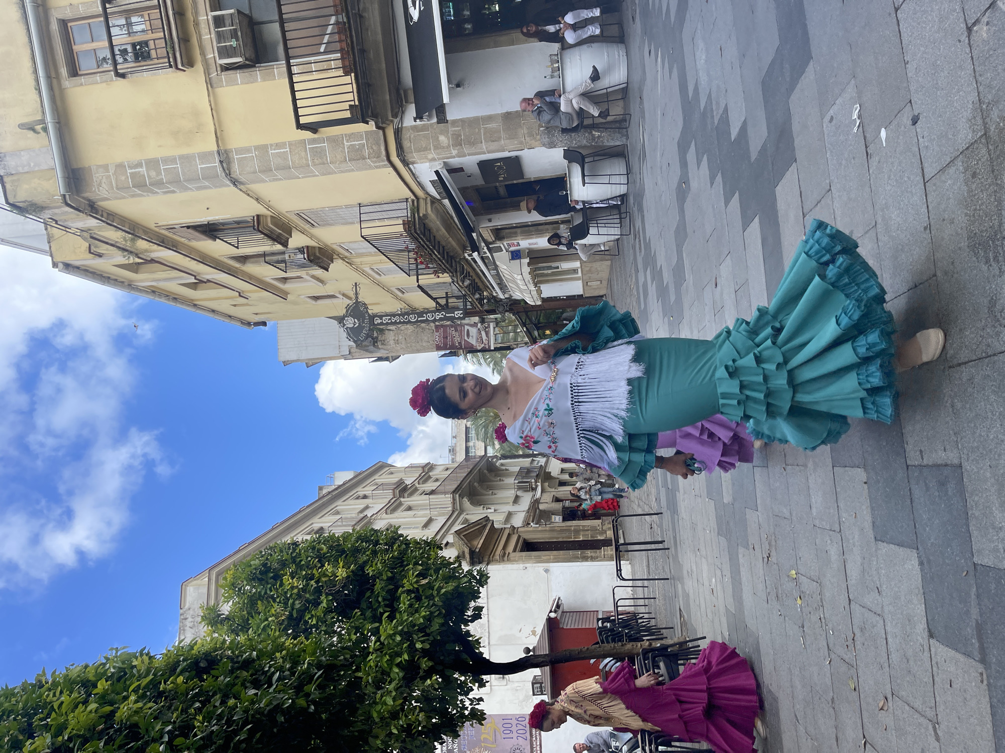 The image depicts a street scene in Jerez de la Frontera, Spain. A woman in a traditional flamenco dress with a large turquoise ruffled skirt and embroidered white top is the central figure. She wears a red flower in her hair. Behind her, there is a building with classical architectural details. Further back, people sit at tables outside a bar or restaurant. Another woman is partially visible in a similar flamenco dress with a purple skirt. A tree is present in the foreground. The ground is paved with grey stone tiles.