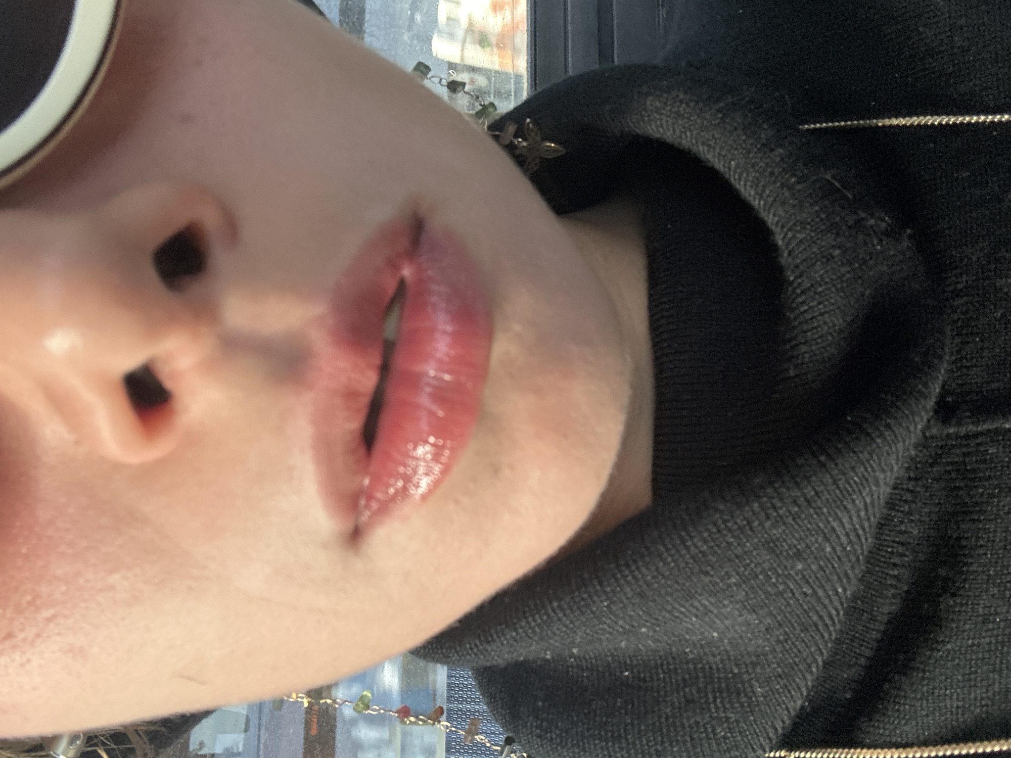 A close-up, sideways-oriented image features the lower part of a person's face, specifically the nose, mouth, and chin, angled towards the top right. The person has a light skin tone. Part of a white-framed sunglass lens is visible at the top left, obscuring the upper face. The lips are parted slightly, revealing a hint of the inner mouth, and are coated in a glossy, pinkish-red lip product. The skin around the nose and mouth appears smooth with some subtle texture.
The person is wearing a black, possibly knitted or ribbed, high-necked garment, likely a sweater or hoodie, which covers the neck and parts of the shoulder, seen on the right side of the frame. In the blurry background, a thin, gold-colored chain with small, multi-colored beads or charms (including green and red) is visible, suggesting a necklace or bracelet. The background also shows indistinct reflections and shapes, possibly from an indoor setting near a window or glass surface, implying a view to the outside or reflections of internal architecture. The lighting appears to be natural daylight. No specific activity is depicted beyond the close-up pose, and no discernible text is visible. The scene is set in what could be an urban environment like Los Angeles, United States of America, though no specific visual cues confirm this.