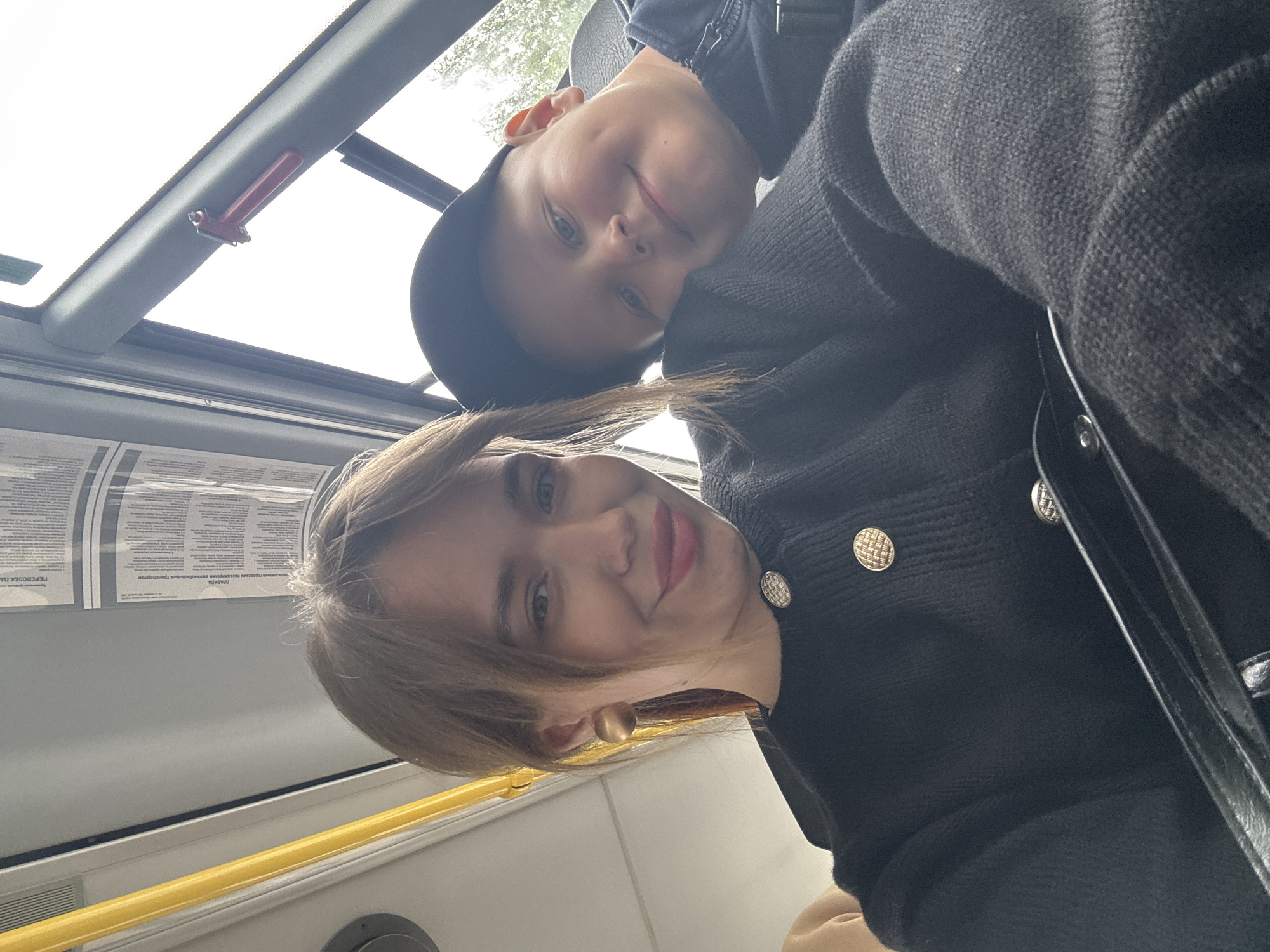 The photo shows a woman and a boy in a bus or trolleybus in Chaikovsky, Russia. The woman has blonde hair and is wearing a black jacket with gold buttons and large round earrings. The boy is wearing a black cap and a dark jacket. Above them is a red emergency hammer on the vehicle's ceiling. A yellow safety bar is visible along the bus wall and promotional posters are placed on the bus windows.