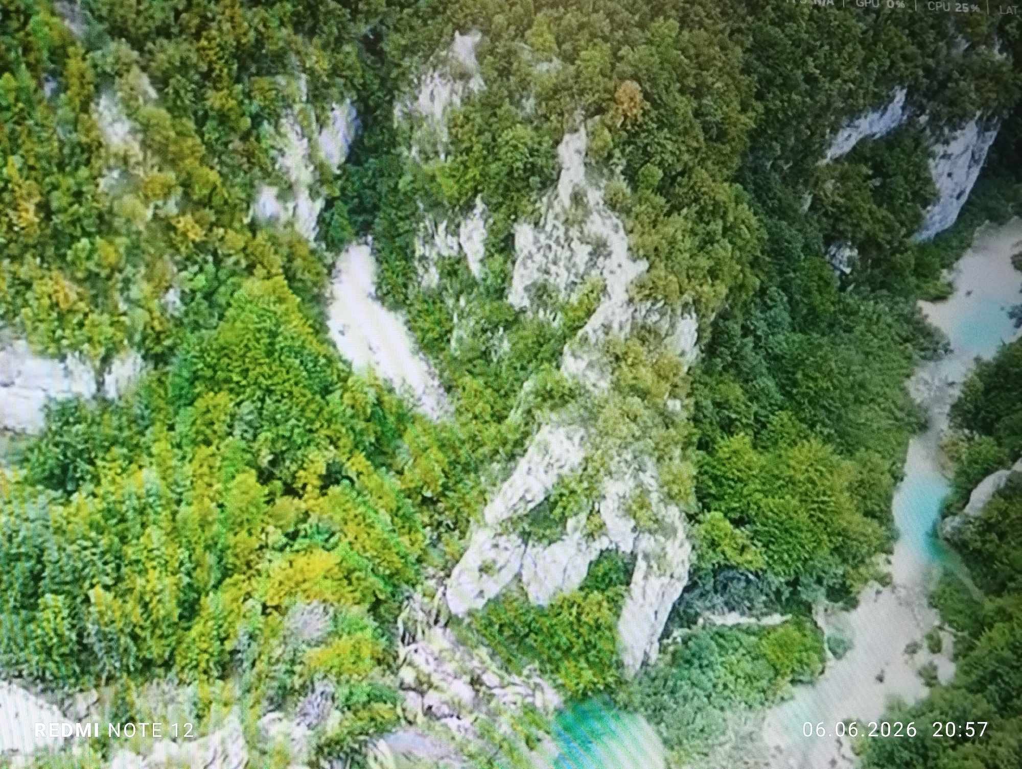 The image displays a high-angle aerial perspective of a natural landscape, dominated by dense green vegetation and numerous exposed white and light grey rock formations. The scene depicts a rugged, possibly mountainous or canyon-like environment.
The majority of the frame is covered by a thick forest, exhibiting various shades of green, from vibrant lime green to darker, more subdued hues. Interspersed throughout the vegetation are prominent, irregular rock outcrops and cliff faces. These rocky areas appear to be limestone or a similar pale geological formation, contrasting sharply with the surrounding foliage.
In the mid-right and extending to the bottom-right of the image, a winding body of water, likely a river or stream, is visible. The water has a distinctive light turquoise or milky blue color, suggesting mineral content or clear, shallow conditions. It flows between vegetated banks and alongside some of the rock formations.
The image itself exhibits noticeable pixelation and a screen-door effect, indicating it is likely a photograph taken of a digital display rather than a direct photograph of the scene. Overlays characteristic of a user interface are present: "FPS N/A | GPU 0% | CPU 25% | LAT" appears in the top-right corner, suggesting the content might be from a game, simulation, or drone telemetry display. A watermark "REDMI NOTE 12" is in the bottom-left, and a timestamp "06.06.2026 20:57" is in the bottom-right, indicating the device and time of the image capture.
No human figures, animals, or man-made structures are visible within the natural landscape depicted. The environment is entirely natural.
While the prompt specifies the location as Burgas, Bulgaria, the image itself does not contain any specific visual identifiers or geographical markers to confirm this exact location within Burgas. The depicted landscape of dense forests, white rock formations, and a turquoise river is characteristic of various natural mountainous or gorge areas globally.