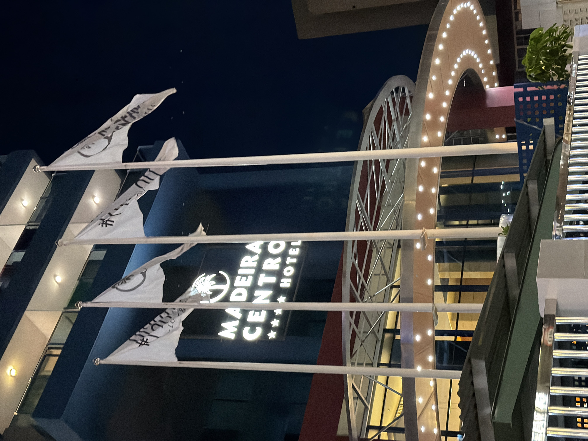 The image shows the exterior of the Madeira Centro Hotel in Benidorm, Spain, at night. The hotel is a tall, dark blue building with illuminated signage that reads "MADEIRA CENTRO ***** HOTEL" with a palm tree logo above the text. Several white flags with black logos are displayed on flagpoles in front of the hotel. The entrance to the hotel features a curved awning illuminated by a string of lights. The scene is set against a dark night sky.