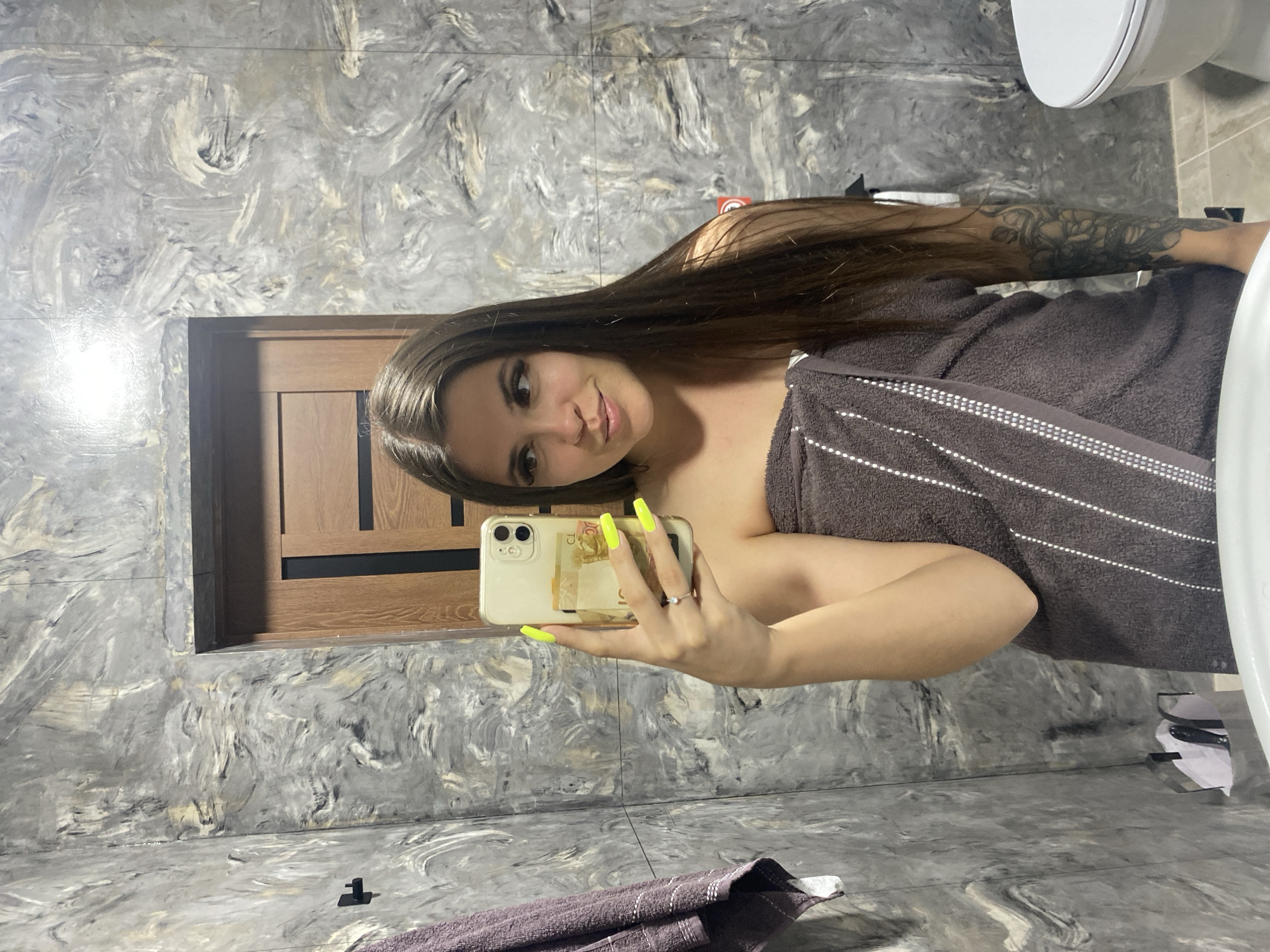 In the image, a woman is taking a mirror selfie in a bathroom. She has long, dark brown hair and is wearing a dark brown towel wrapped around her body. Her nails are painted a bright neon yellow. She is wearing a ring on her left ring finger. Behind her, a wooden door with black accents is visible. The bathroom walls have a grey, marbled texture. A purple towel hangs on a hook to the left of the woman. To the right, a toilet with a white lid is partially visible. The image appears to be taken in a residential bathroom, potentially in Chalda, Russia.