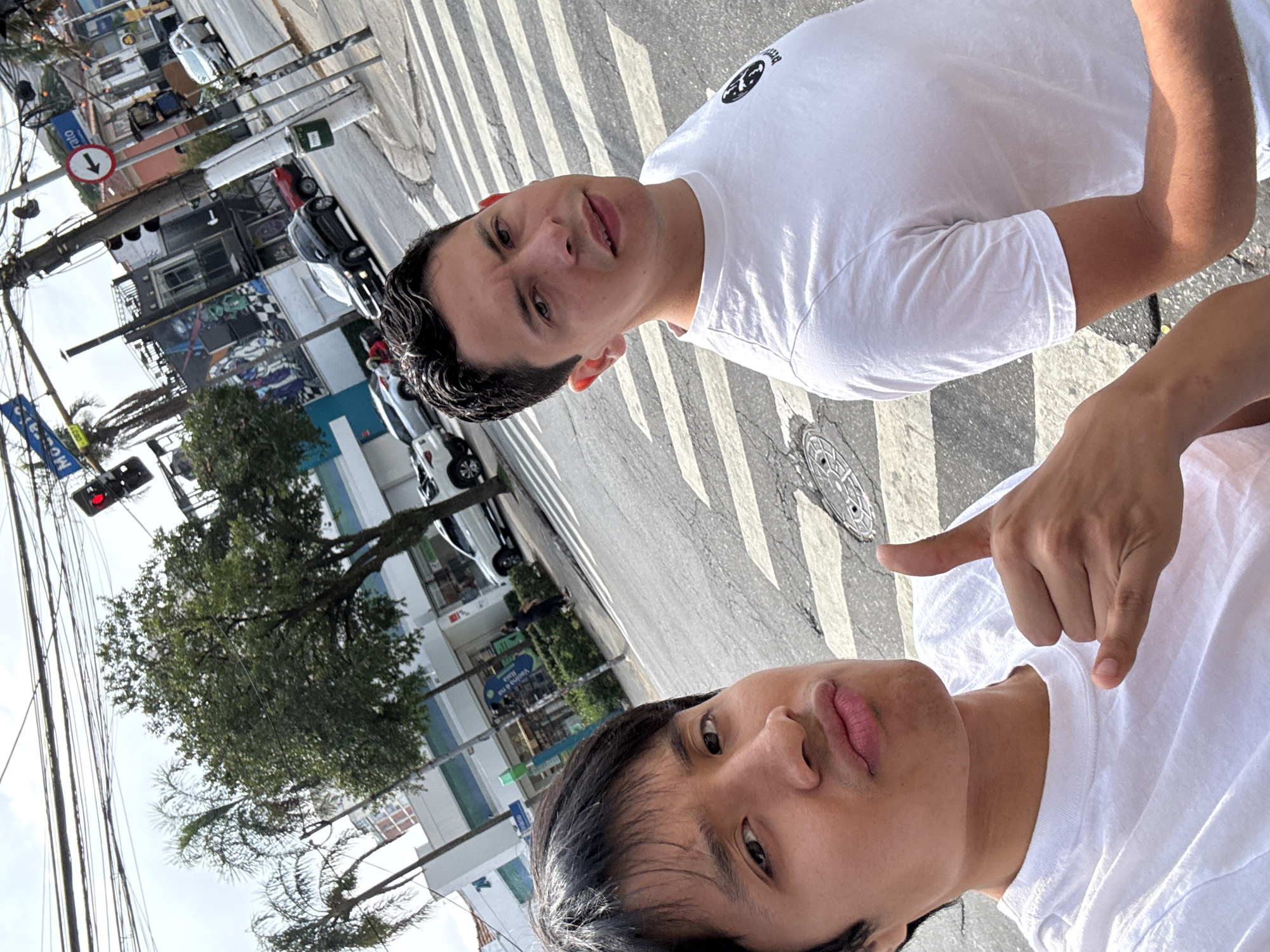 The image captures two young men taking a selfie on an urban street in São Paulo, Brazil, under bright daylight. The scene is rotated, making the subjects and background appear as if viewed from an unusual angle.
In the foreground, one man with dark hair, wearing a white t-shirt, looks towards the camera with a slight pout on his lips. His right hand is raised, making a pointing-down gesture. Partially obscured behind him, another man with dark, slicked-back hair, also in a white t-shirt, is fully visible, looking at the camera with a subtle smile. A dark, circular logo is visible on the chest of his t-shirt.
The background depicts a vibrant street scene. The asphalt is marked with clear zebra stripes of a pedestrian crossing, and distinct shadows indicate bright, sunny weather. Buildings line the street, some featuring graffiti-style art on their lower facades. Various cars are parked along the curb and appear in the distance. To the left, tall utility poles are laden with a dense network of overhead wires. A traffic light displays a red signal. Lush green trees are scattered throughout the scene.
Several pieces of text and notable signs are visible: a blue street sign on a pole reads "MORAES," another blue sign further back possibly says "Valinhos," and a white circular sign with a red border and a white downward arrow, indicating a one-way street, is also present in the mid-ground. A manhole cover is visible on the street surface. The overall setting is that of a lively city environment.