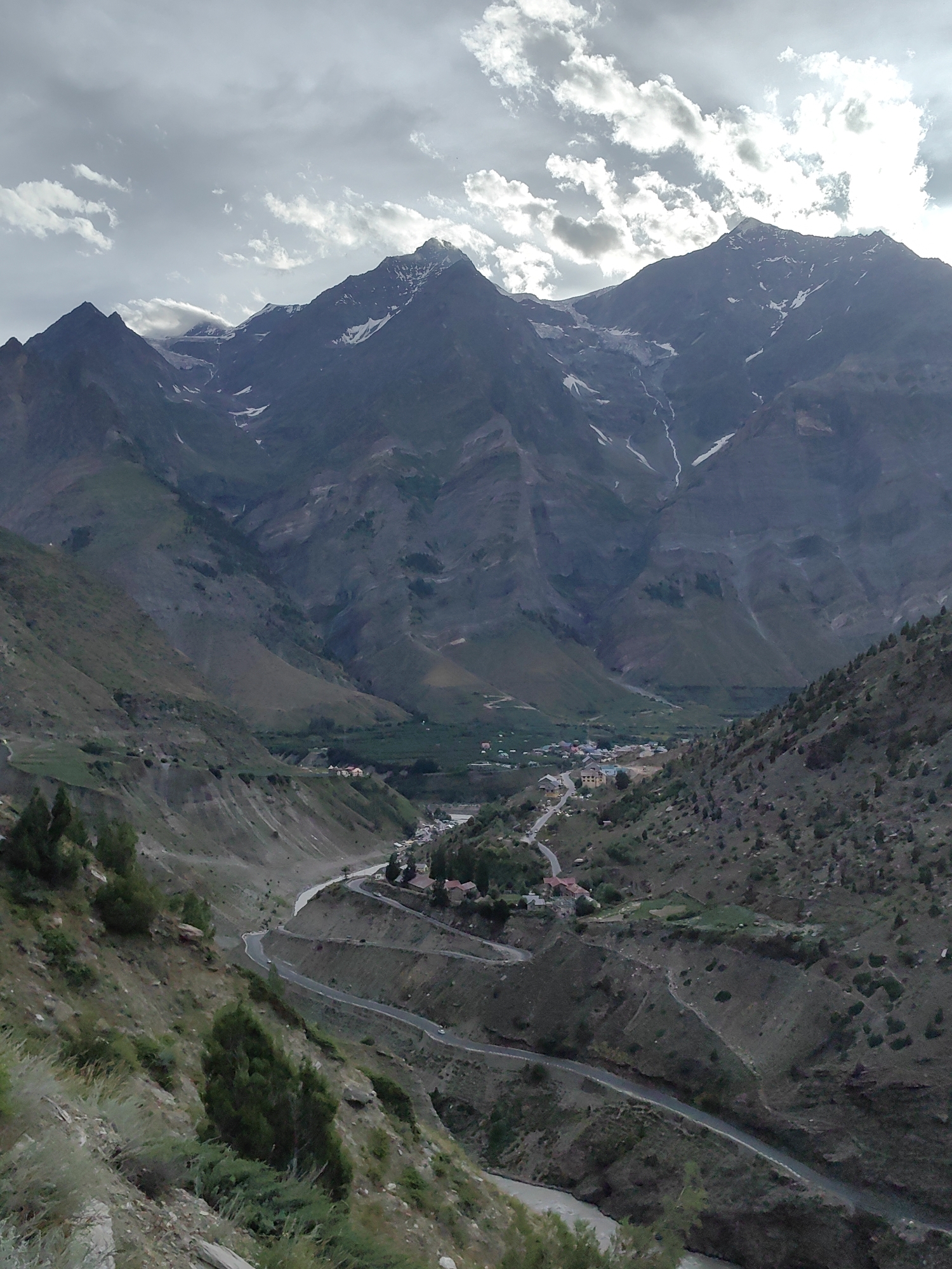 A vertical, high-angle, wide shot captures a mountain valley in Tandi, India, under a cloudy sky. The foreground shows a steep, rocky slope covered in sparse vegetation, including grasses and bushes. A winding road snakes its way down the slope and into the valley. In the middle ground, a small cluster of buildings, appearing to be homes or a village, are nestled on the valley floor. Further in the distance, a river flows through the valley. The background is dominated by a massive mountain range with jagged peaks. Some of these peaks are snow-capped, and streaks of snow are visible on the slopes. The mountains are rugged and layered, with visible rock formations and vegetation clinging to their surfaces. The overall scene conveys a sense of remote, natural beauty and a small human settlement existing within a vast mountainous landscape.