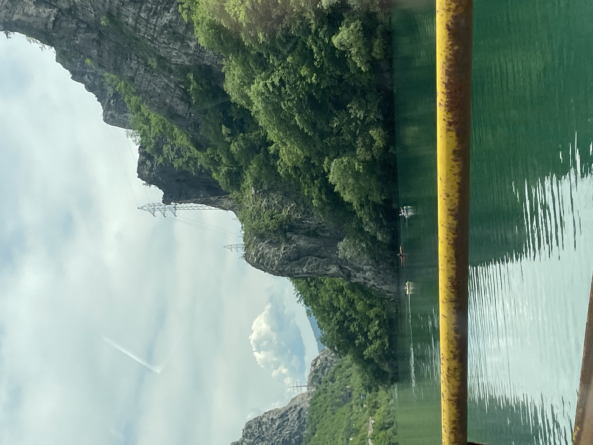 The image captures a scene on the water near Kremenac, Bosnia and Herzegovina. In the foreground, a yellow metal pole frames the view. Beyond the pole, the water reflects the sky, displaying a mix of light and darker shades. On the left side, a rocky mountain covered with lush green trees rises. Power lines are visible extending across the landscape, connecting to metal towers perched on the mountain. Two people in kayaks are visible on the water near the shore. The sky is overcast with clouds.