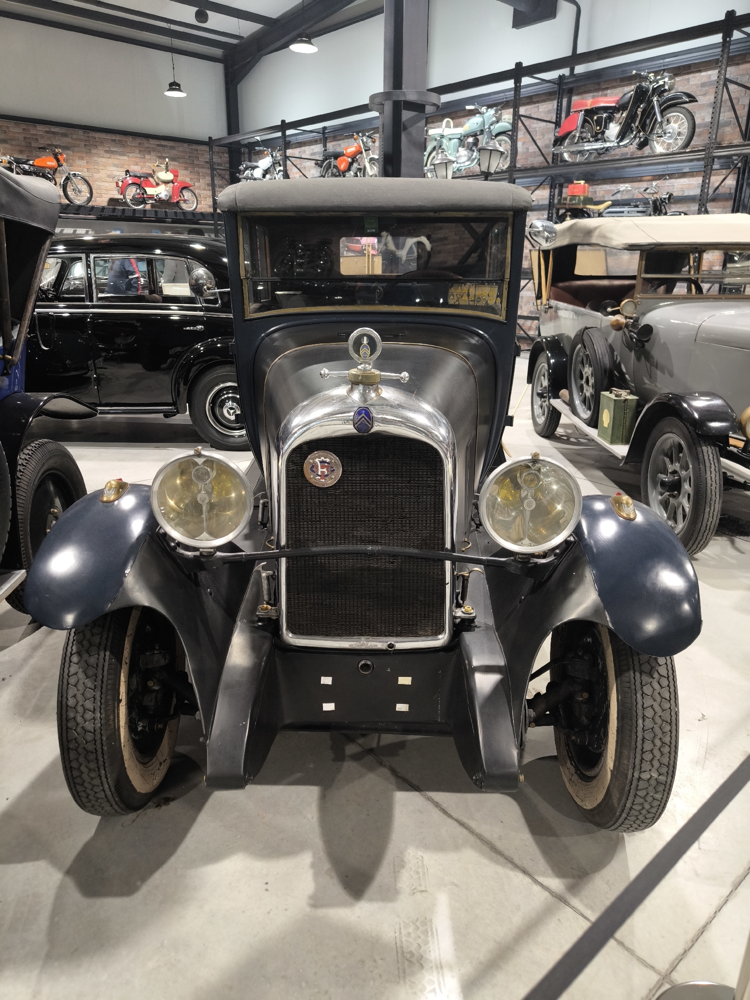 The image shows a collection of vintage vehicles housed inside a building with a high ceiling and a brick wall in the background. The foreground is dominated by a dark blue vintage car with large headlights and a prominent grill. To the left, a black sedan is partially visible. To the right, another grey vintage car is present. On shelves along the brick wall, several vintage motorcycles are displayed. The environment appears to be a museum or private collection. The location, Kapatovo, Bulgaria, is not evident from the image.