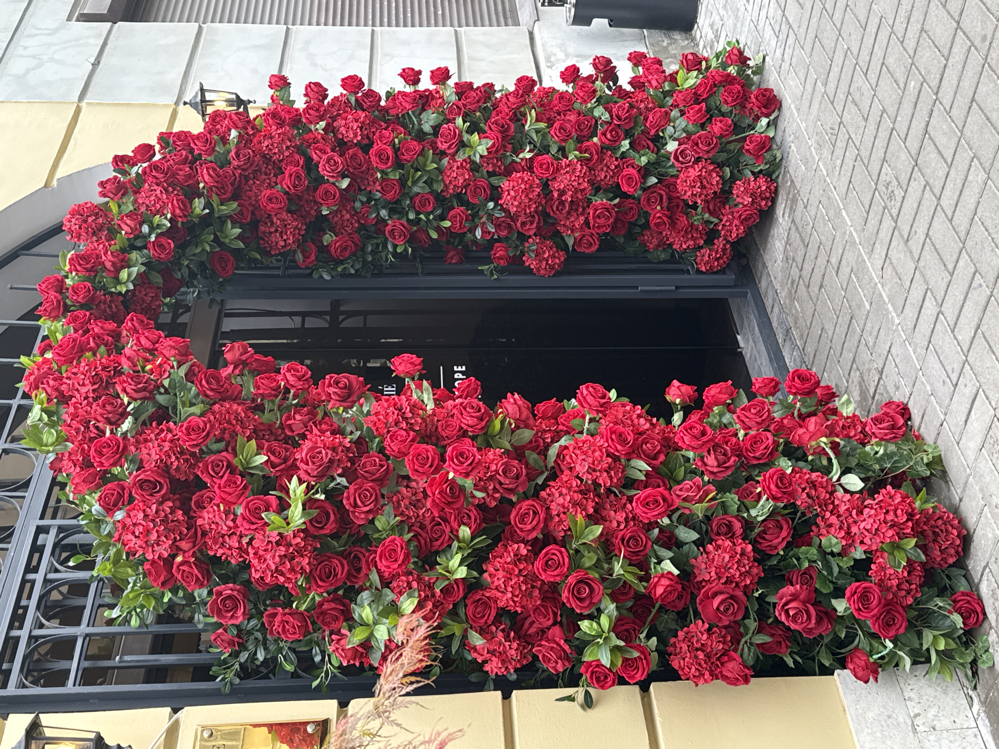 The image shows a shop entrance heavily decorated with red roses and hydrangeas. The floral arrangement frames the doorway in a half-arch shape above and a more filled-in arrangement below. The building appears to have a grey brick facade to the right of the doorway and a yellow painted facade to the left. A black metal grid is visible on the left side of the floral arrangement. Based on the decor and architecture, it is possible that this photo was taken in Saint Petersburg, Russia, although there are no definitive markers in the image to confirm this.