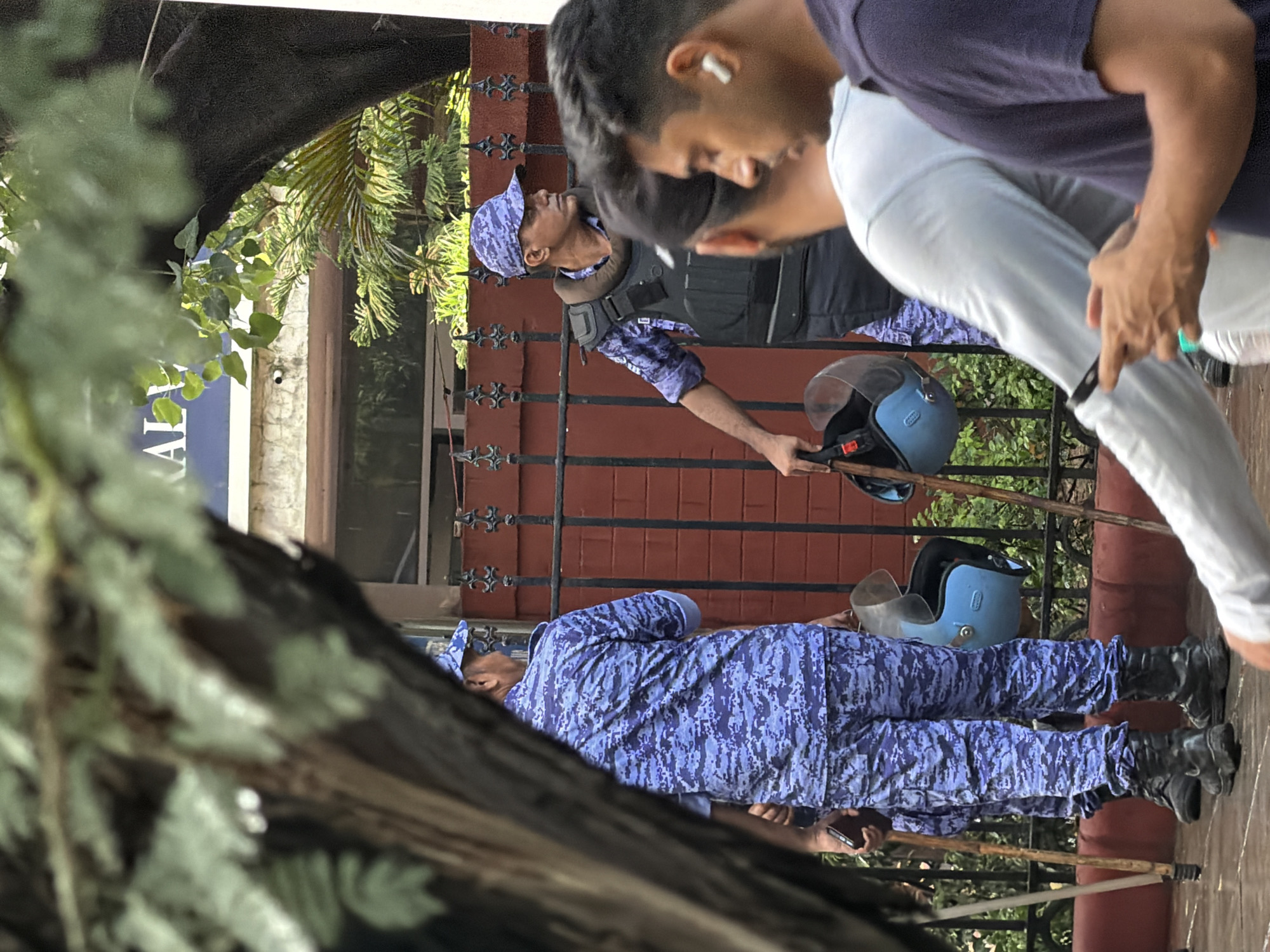 In New Delhi, India, a contingent of police officers in camouflage uniforms and protective vests stand near a red brick wall and a black metal fence. One officer holds a helmet, while another is in the process of putting on a protective vest. Several individuals are visible in the foreground, including a man in a light blue shirt holding a mobile phone. The background reveals a building with a sign that appears to read "RALY" and trees with lush green foliage. The scene suggests a security presence or a public event in an urban setting.